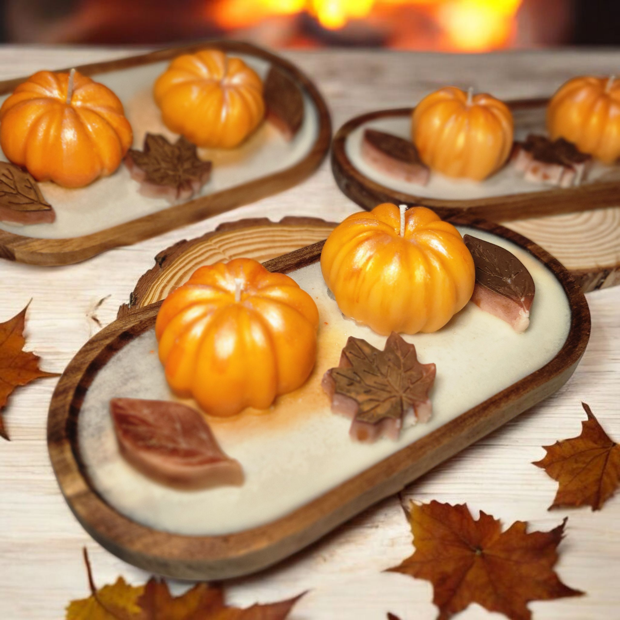 Decorative pumpkin candles on wooden trays, surrounded by autumn leaves, with a blurred warm-colored background.