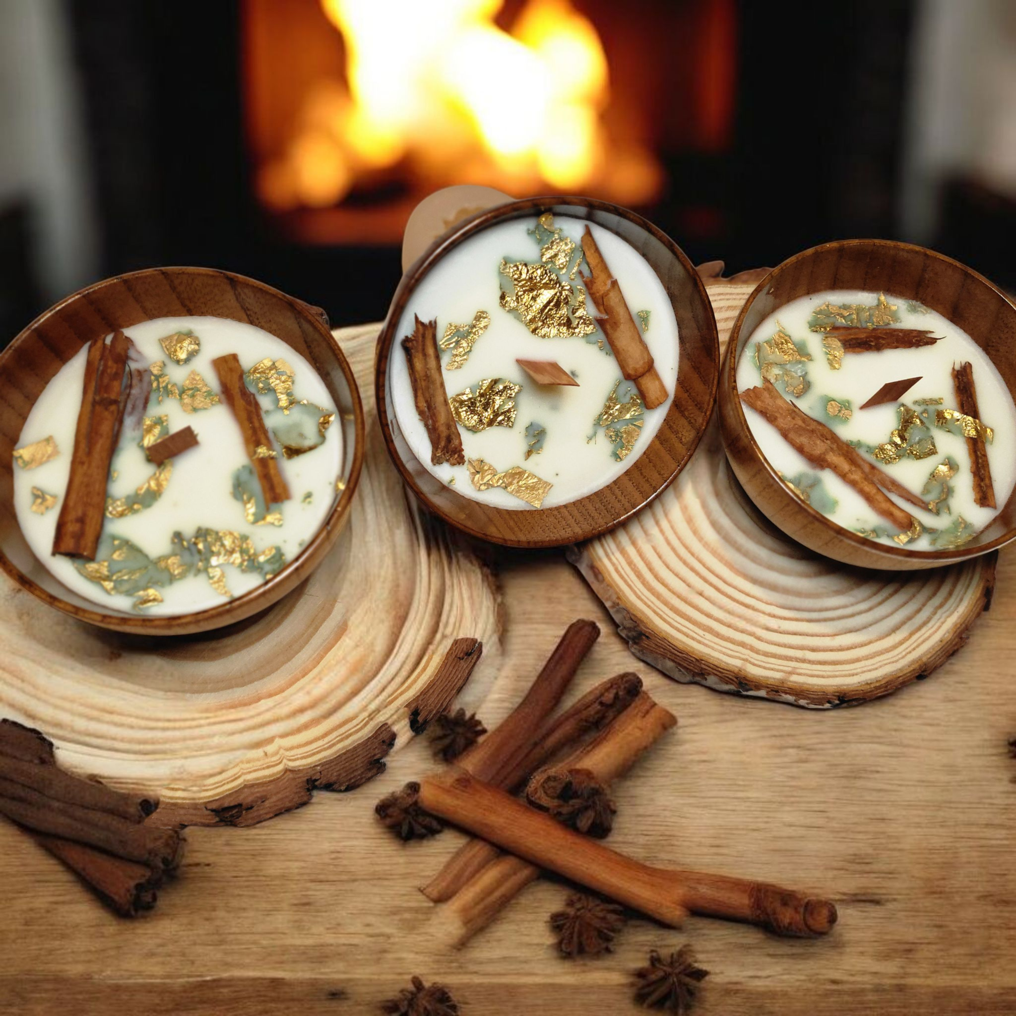 Three lit candles in wooden holders on a wooden surface, with cinnamon sticks and star anise in front, flickering in front of a fireplace.