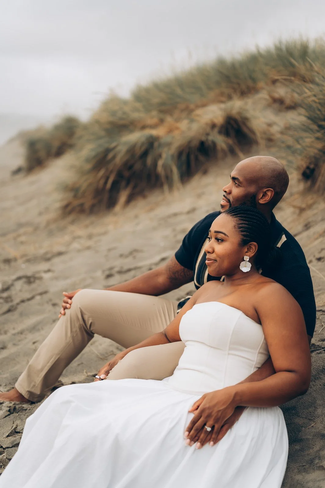 A couple sitting on the beach, the woman in a white dress and the man in a dark shirt, with sand dunes and grasses in the background.