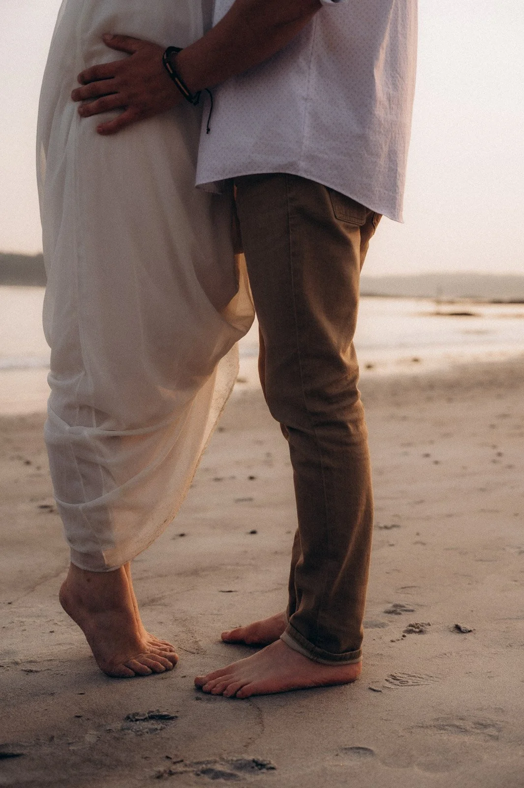 A couple standing on the beach, holding hands, with their feet in the sand during sunset.