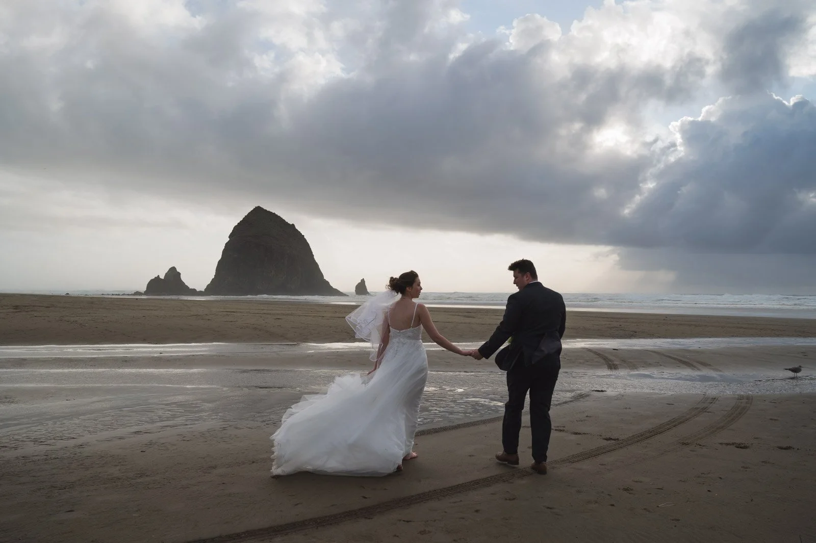 A bride and groom walk hand in hand at Cannon Beach with overcast sky and Haystack Rock in the background.