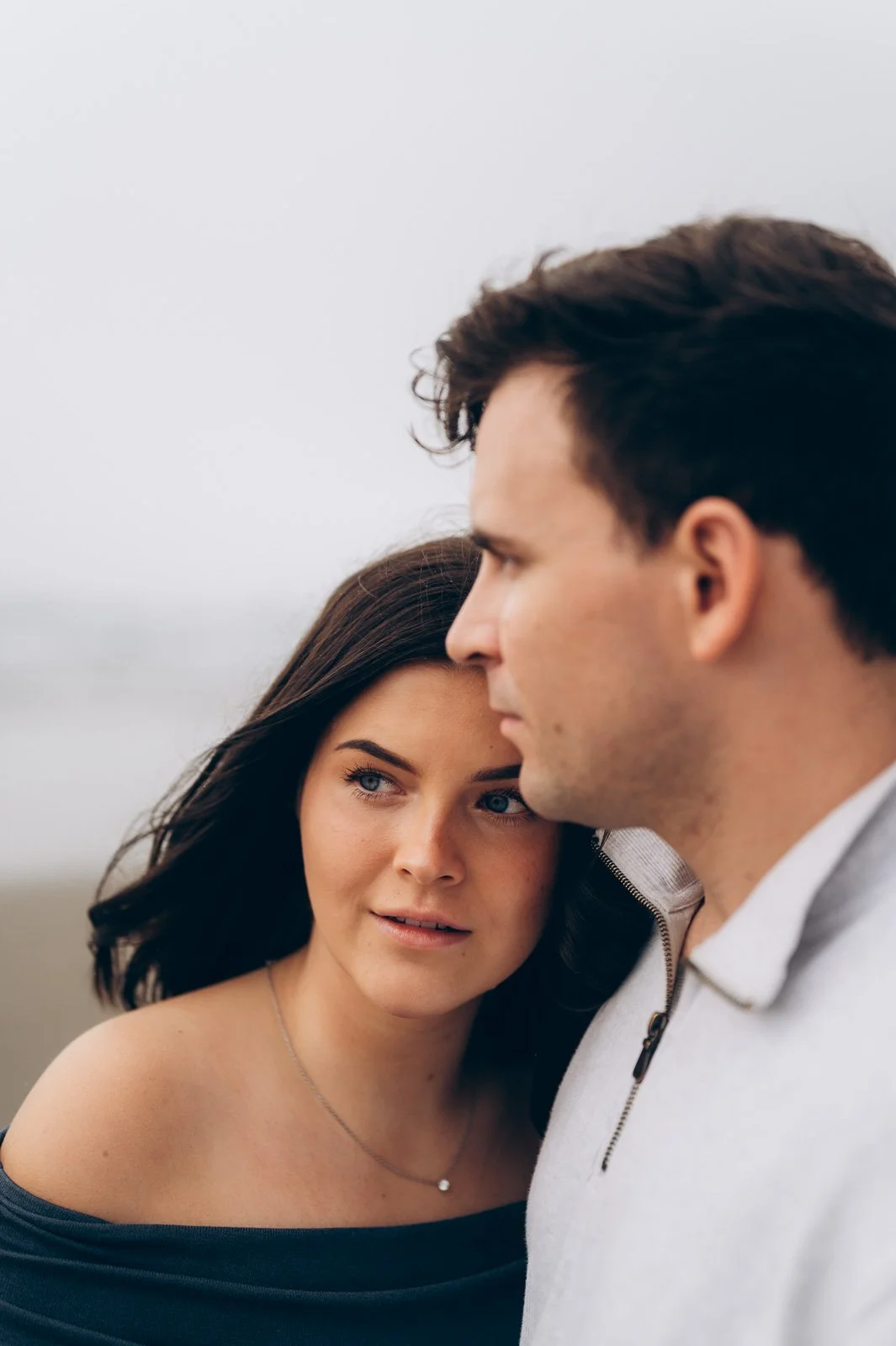 A woman and a man standing close together outdoors, with the woman resting her head on the man's shoulder.