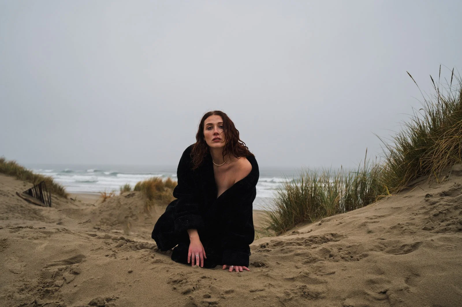 A woman with curly brown hair, wearing a black coat, sitting on sandy dunes with the ocean and overcast sky in the background.