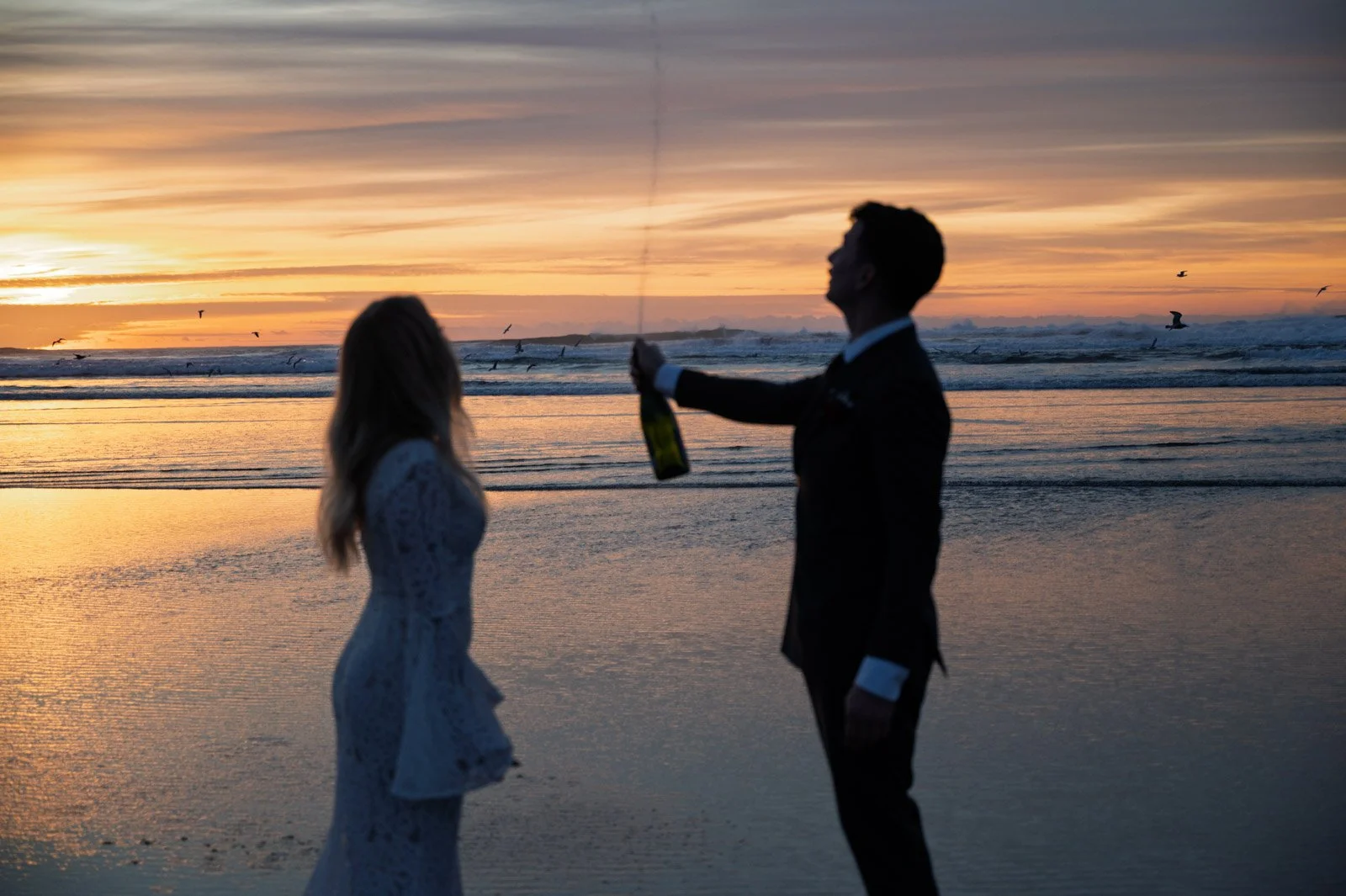 A silhouette of a man in a tuxedo pouring champagne for a woman in a white lace dress on the beach at sunset, with birds flying in the sky.