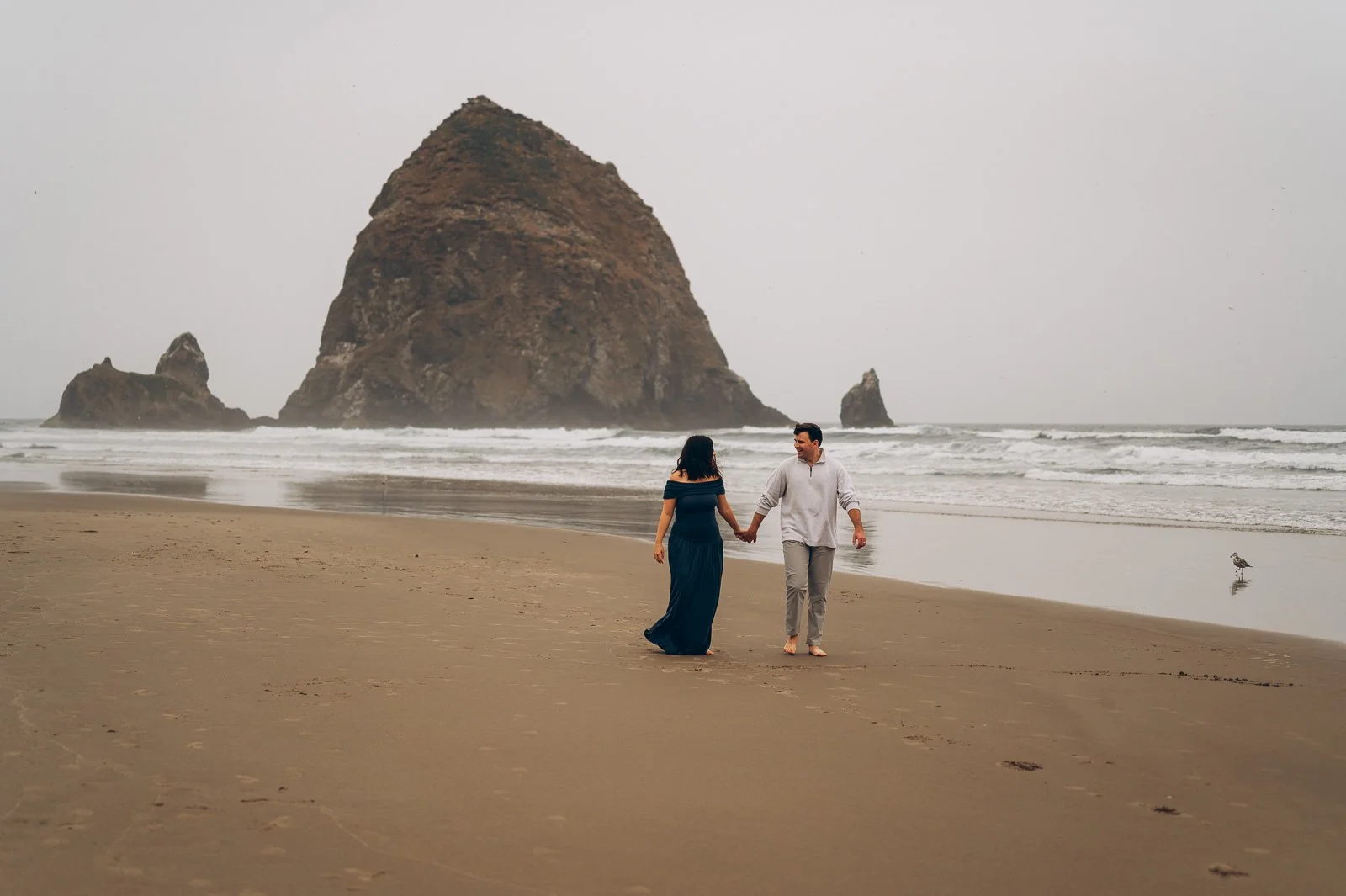 A couple walking hand in hand on Cannon Beach with Haystack Rock in the background and a cloudy sky.
