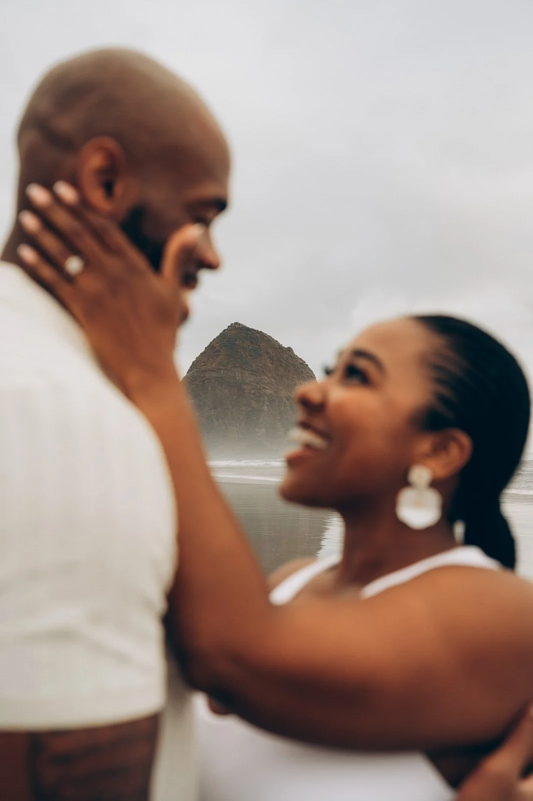 A couple on Cannon Beach with Haystack Rock in the background, smiling and looking at each other. The man has a shaved head and a beard, the woman has dark hair pulled back and is wearing earrings.