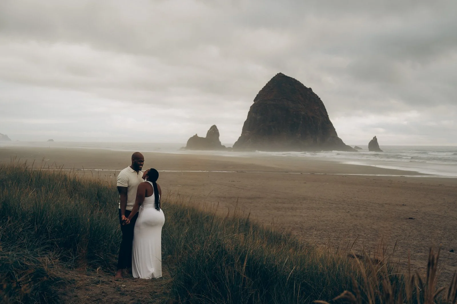 A couple holding hands at Cannon Beach, with Haystack Rock and an overcast sky in the background.