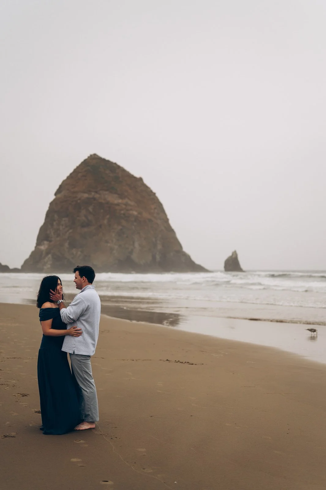 A couple standing at Cannon Beach holding each other, with Haystack Rock in the background and the ocean waves.