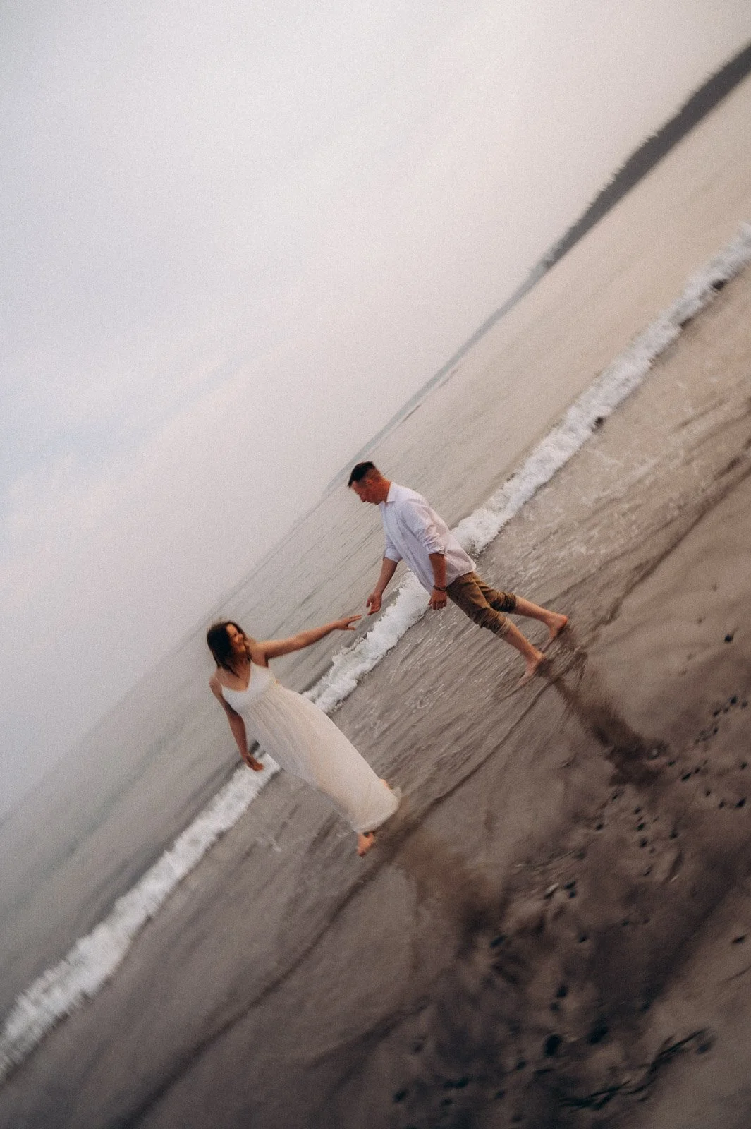 A woman in a white dress and a man in a white shirt and khaki pants holding hands in the ocean surf on the beach during sunset.
