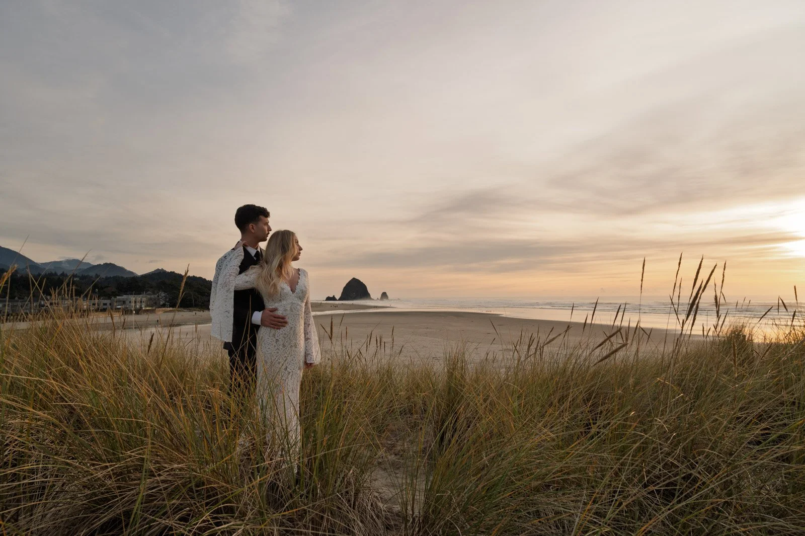 A couple dressed in wedding attire standing on Cannon Beach during sunset, with grassy dunes in the foreground and rock formations in the distance.