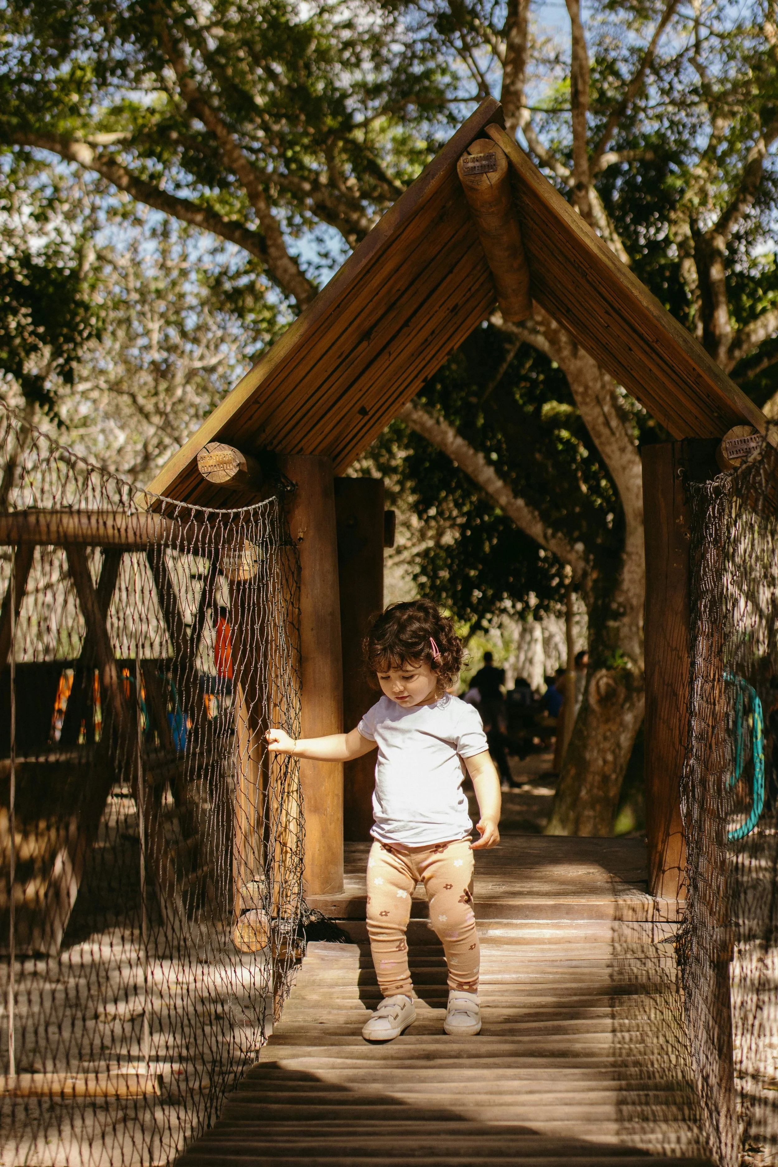 A young child with curly hair, wearing a white shirt and patterned leggings, standing on a wooden bridge with a fence on each side, under a wooden structure with a triangular roof, surrounded by trees.