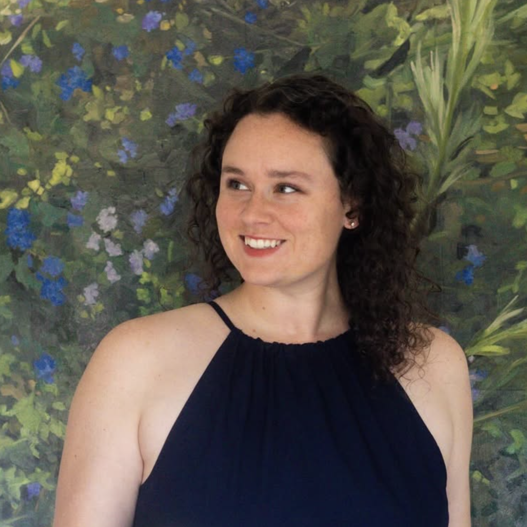 A woman with curly dark hair wearing a navy blue sleeveless top standing in front of a floral background with green leaves and blue flowers. Kacie Landis