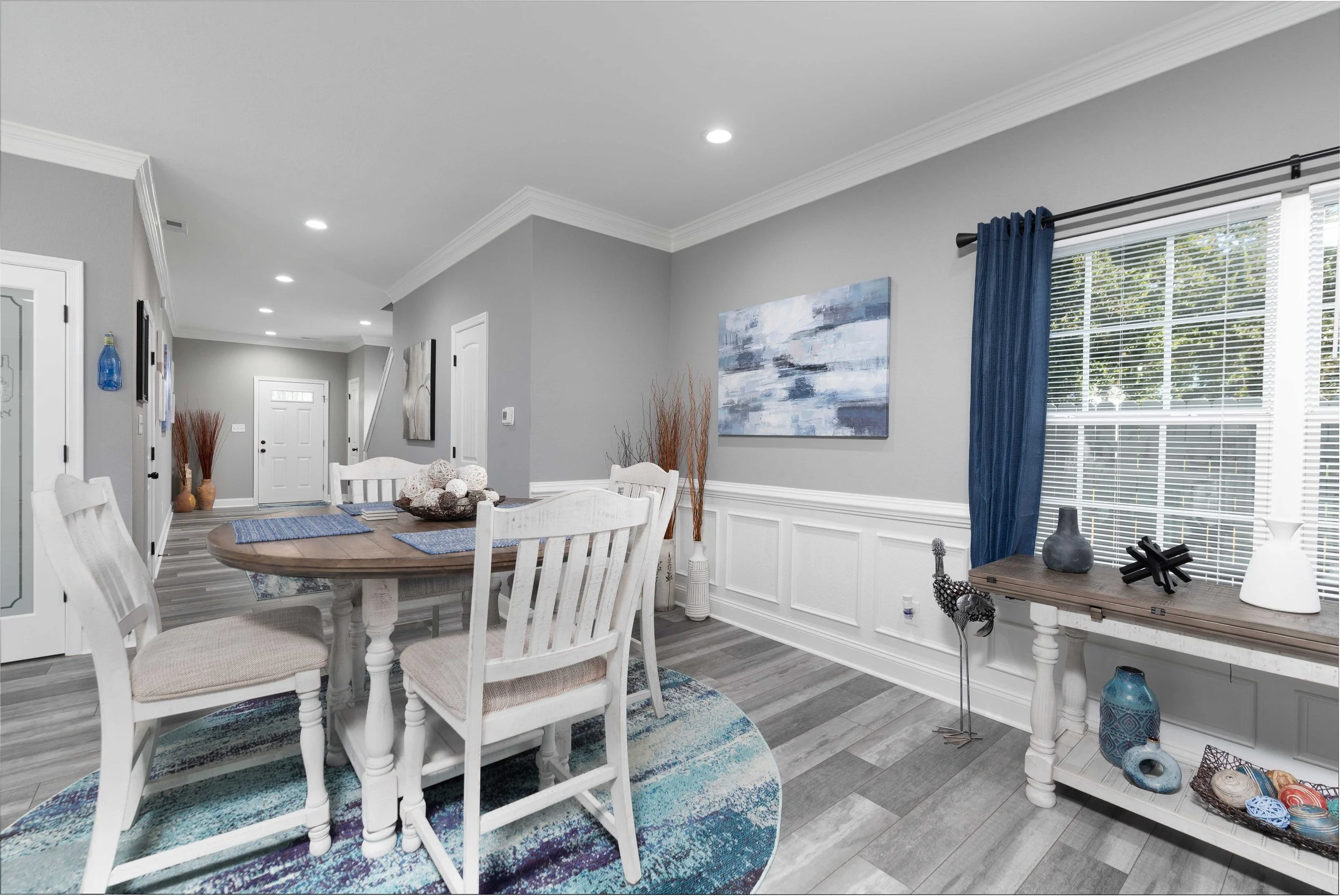 Living and dining room area with gray walls, white wainscoting, a round wooden dining table with four white chairs, a blue curtain on a window, a wooden console table with decorative vases and sculptures, a colorful rug, and abstract paintings on the walls.