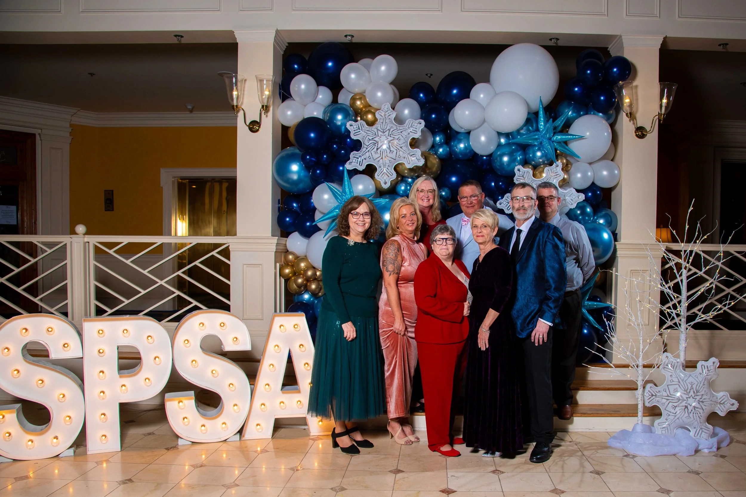 A group of people posing in front of a festive holiday backdrop with blue, white, and gold balloons, snowflake decorations, and large lit letters spelling 'SPSA' at the front.