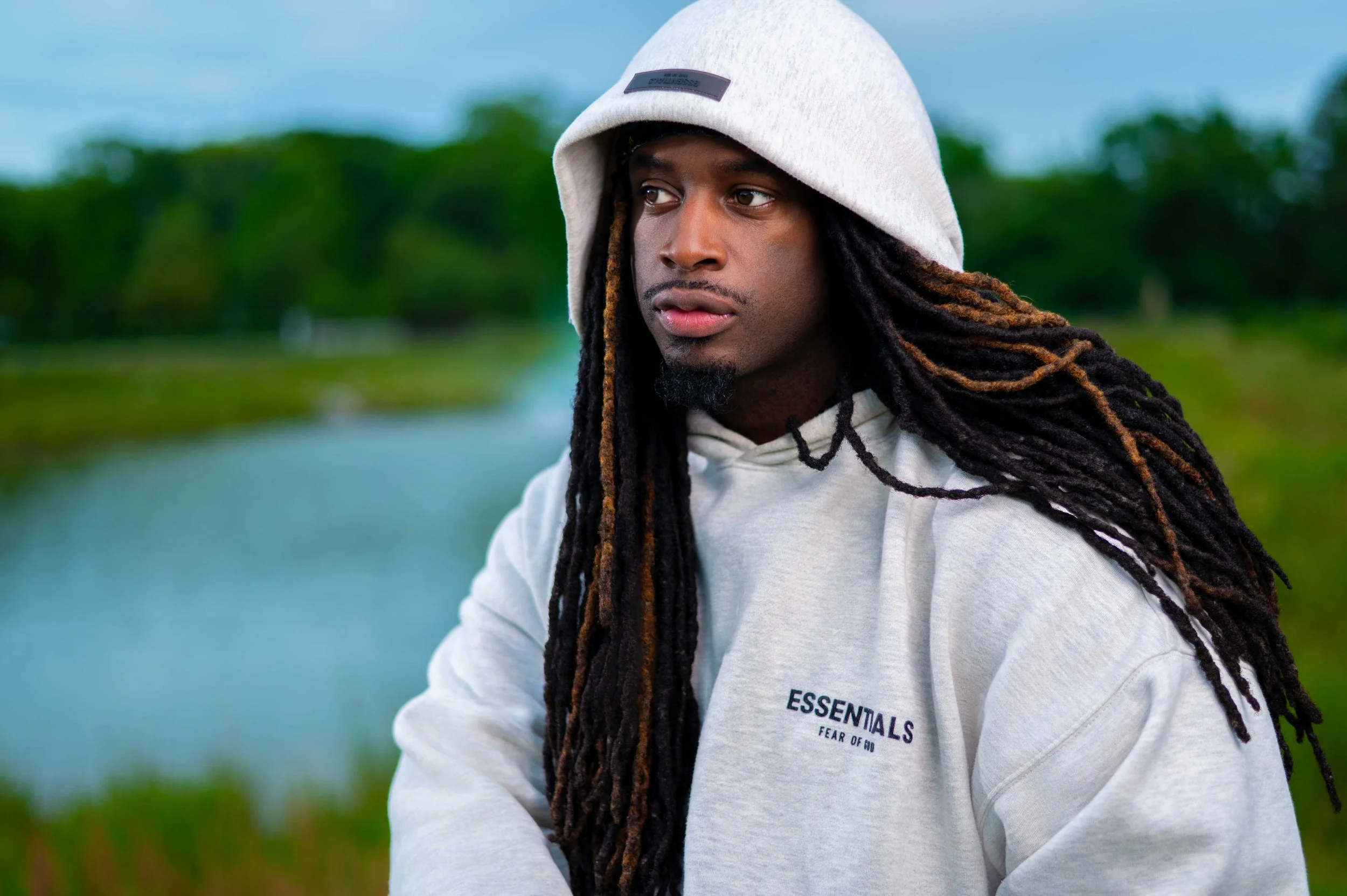 A young man with long dreadlocks wearing a white hoodie and beanie, standing outdoors near a body of water with greenery in the background.