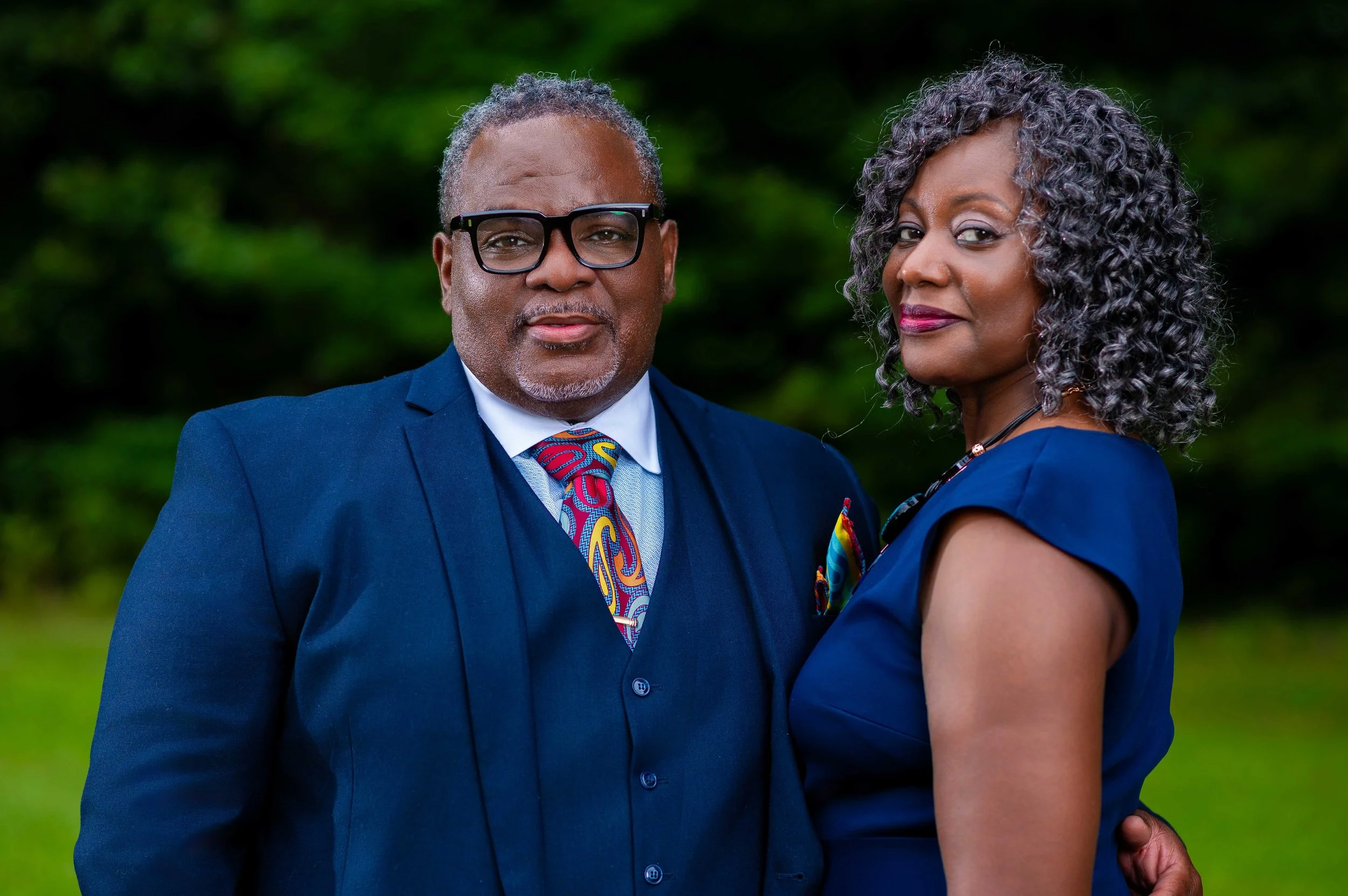 A mature African American man and woman standing outdoors with a blurred green background. The man is wearing a navy blue suit, a colorful tie, a white shirt, black-rimmed glasses, and has gray hair. The woman is dressed in a navy blue dress, has curly gray hair, and is wearing makeup with a focus on her lips.