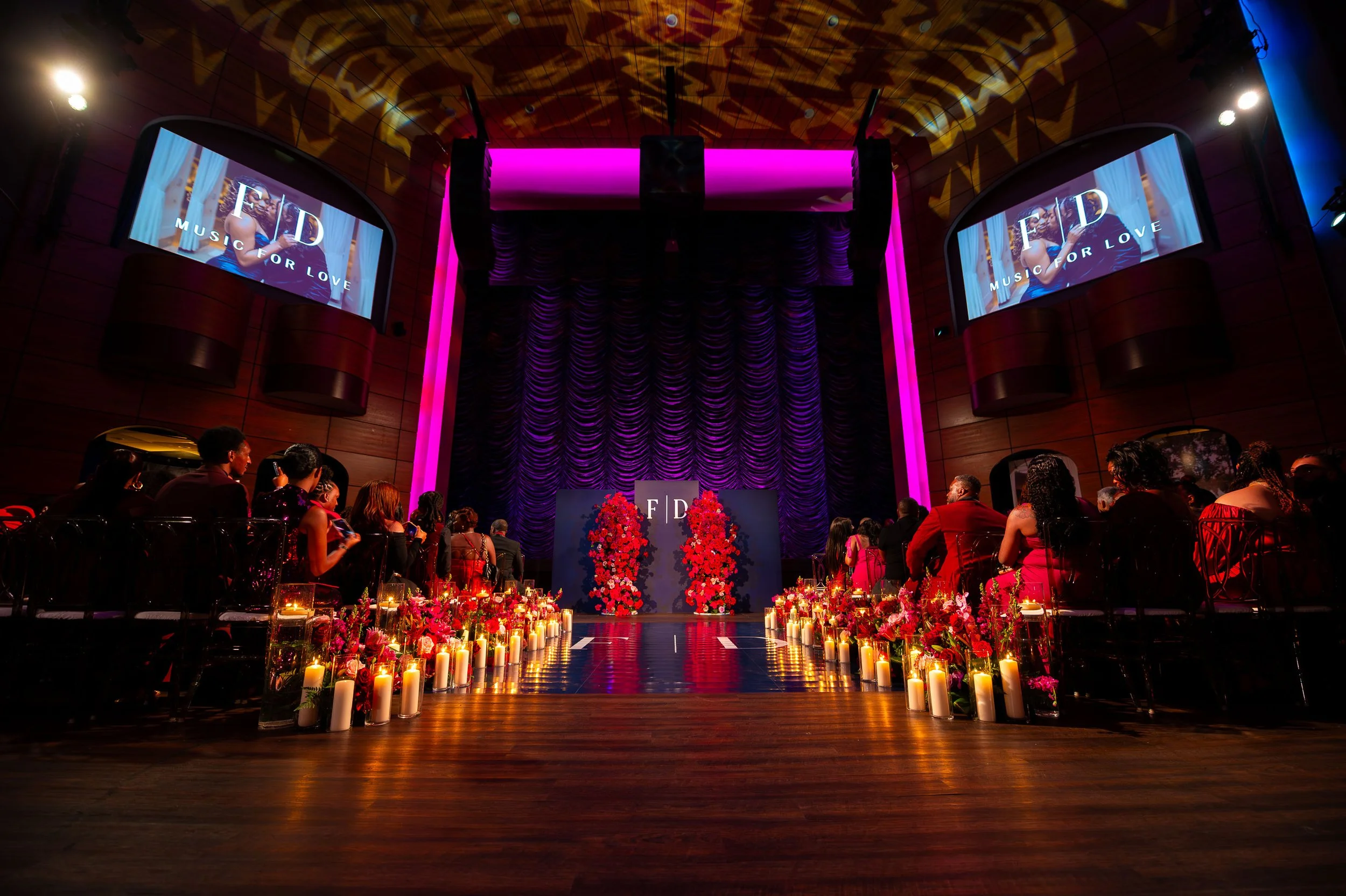 An indoor event with a large stage decorated with purple curtains and two large floral arrangements of red roses. The stage has a sign with the initials 'F D'. Audience members dressed in formal attire are seated along a decorated aisle lined with candles and flowers. Two large screens display the logo 'F D' and the phrase 'Music for Love'.