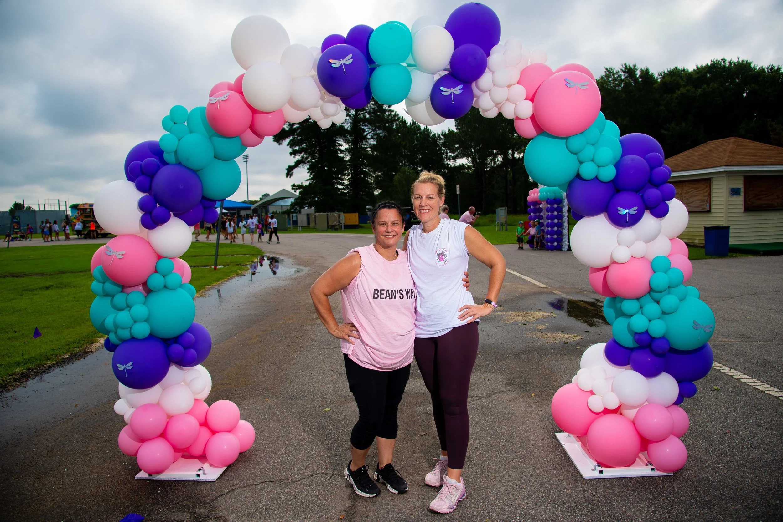 Two women standing together under a colorful balloon arch at an outdoor event, with other people and structures in the background.