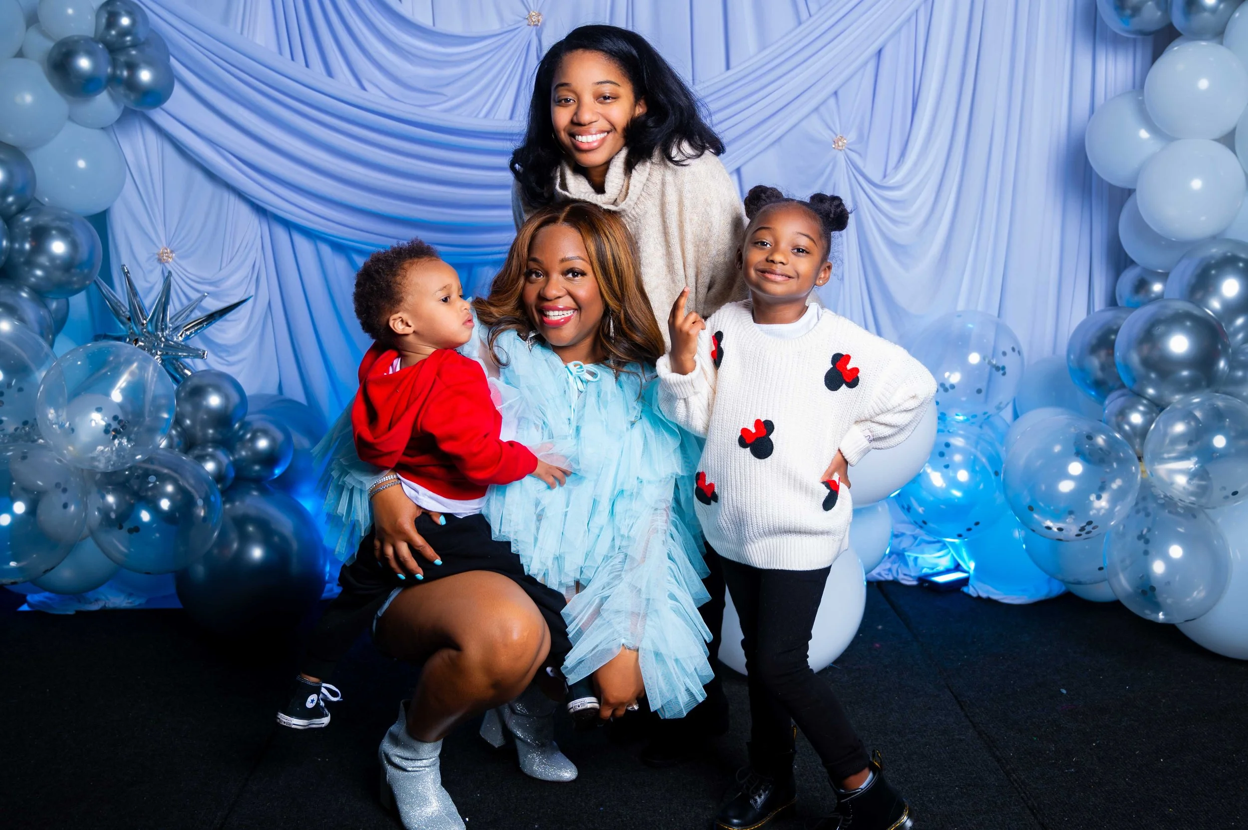 A woman and three children pose for a photo in front of a blue curtain decorated with silver and white balloons, some transparent with dark confetti inside. The woman is sitting on her knees, wearing a light blue outfit, and smiling. One girl, standing to her right, is dressed in a white sweater with red and black Mickey Mouse designs and black pants, pointing up with her finger. Another girl, standing behind the woman, has curly hair, a beige sweater, and is smiling. The third child, a boy, is wearing a red hoodie and black shorts, sitting on the woman's lap and looking at the girl in the sweater.