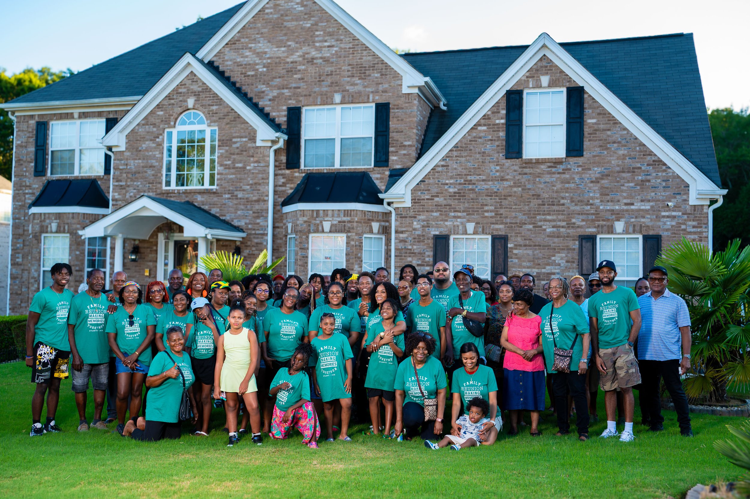 A large group of people gathered in front of a two-story brick house with green grass and palm plants around them. Many are wearing matching teal T-shirts, and there are children and adults of various ages smiling for the photo.