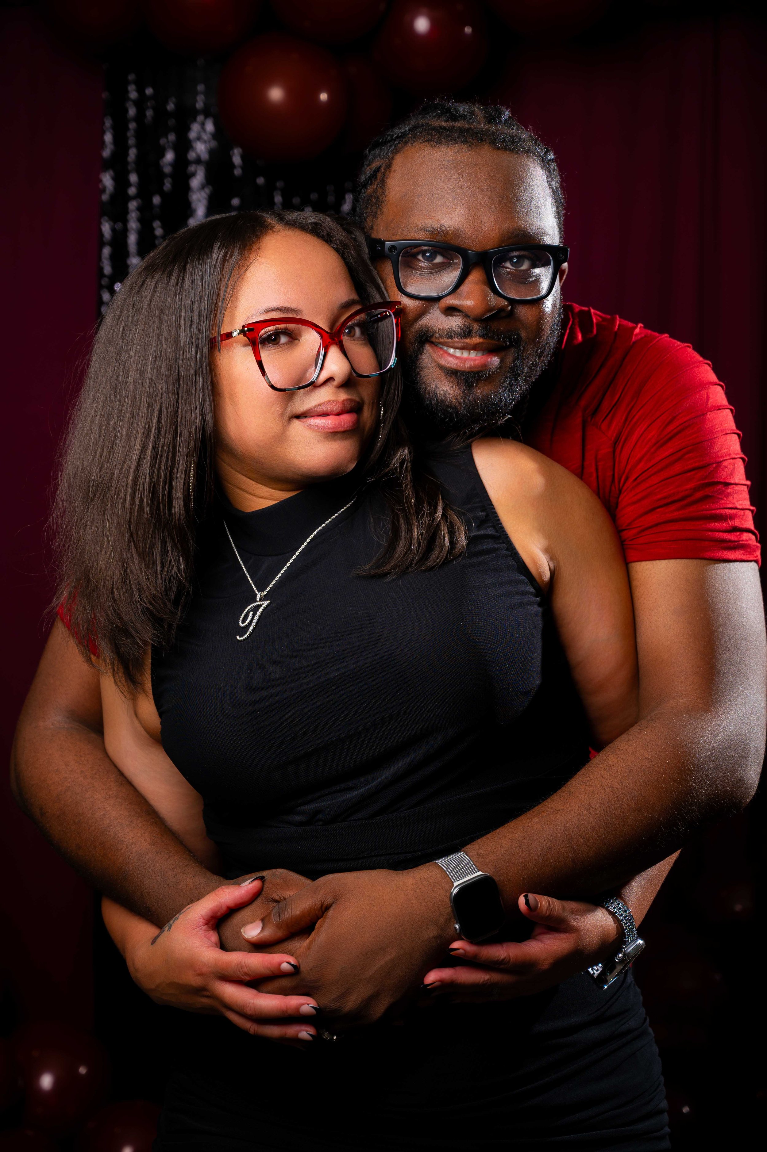 A couple embracing at a party, with the woman wearing glasses, a black sleeveless dress, and a necklace, and the man wearing glasses and a red shirt, standing close together against a decorated dark background with balloons.
