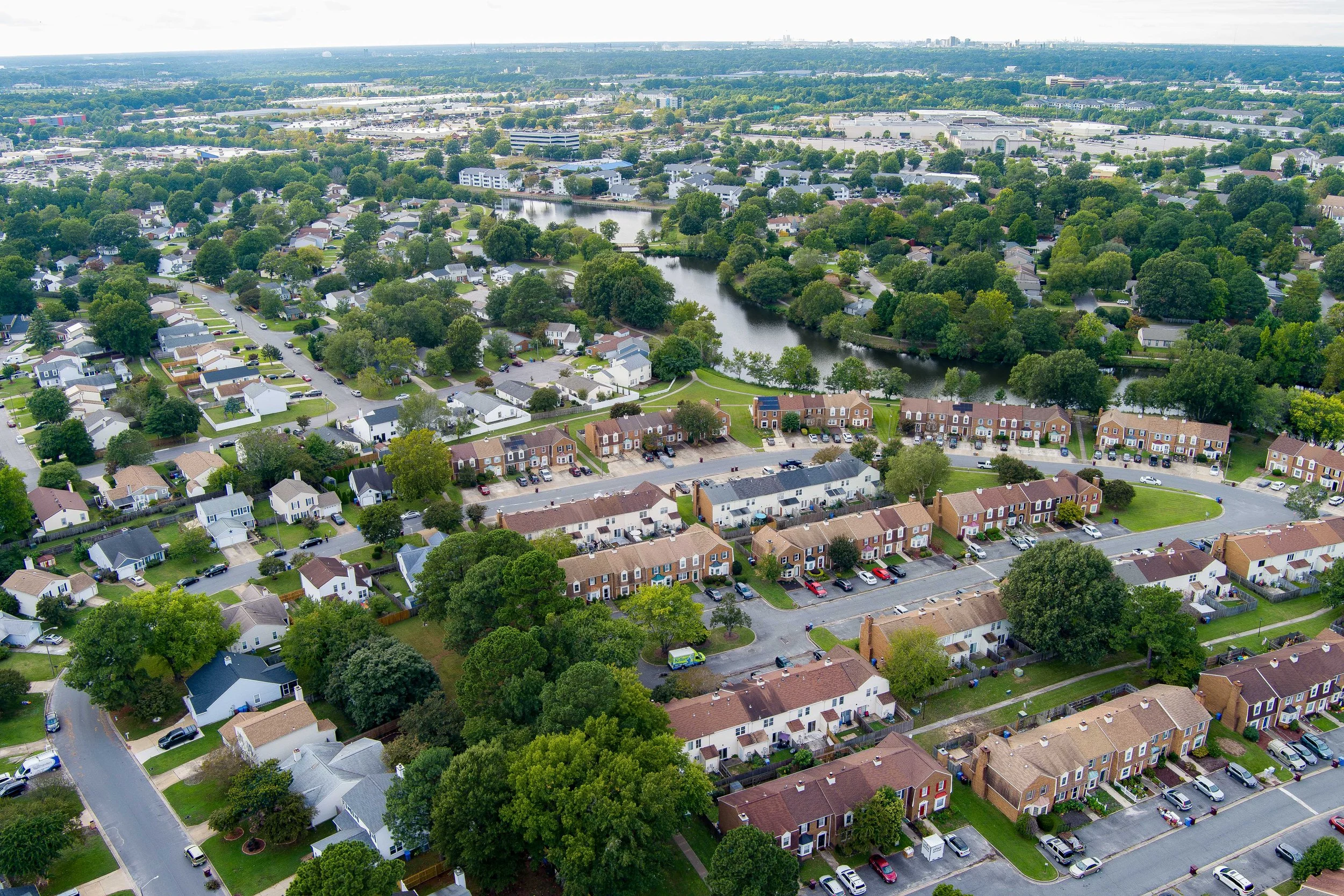 An aerial view of a suburban neighborhood with houses, trees, a pond, and parking lots, with a cityscape in the background.