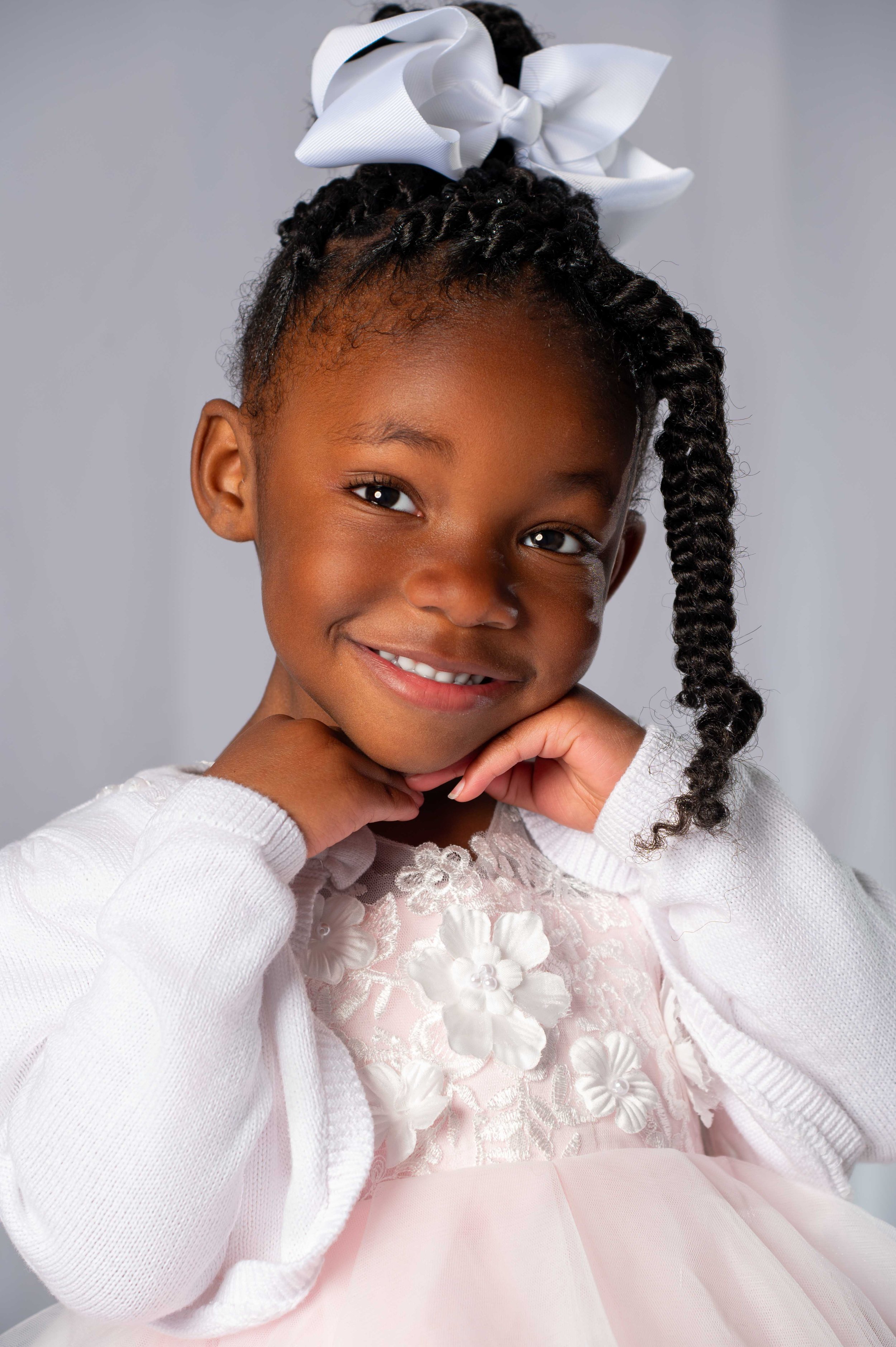 A young girl with braided hair and a big white bow, smiling and resting her chin on her hands, dressed in a light pink dress with floral embroidery and a white cardigan.