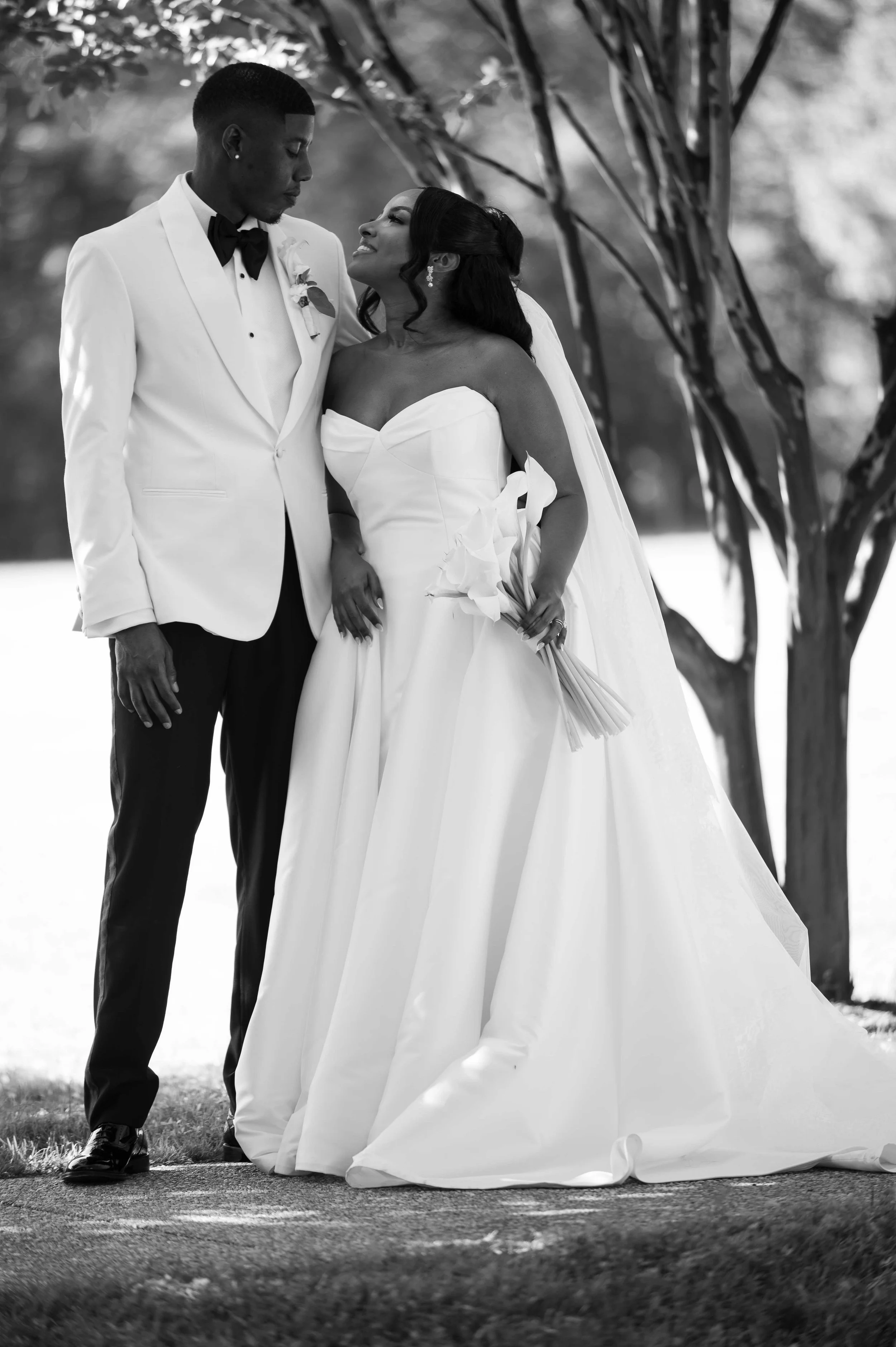 Black and white photo of a bride and groom standing outdoors near a tree, looking at each other. The groom wears a white tuxedo with a black bow tie, while the bride wears a strapless white wedding gown and holds a bouquet of calla lilies.