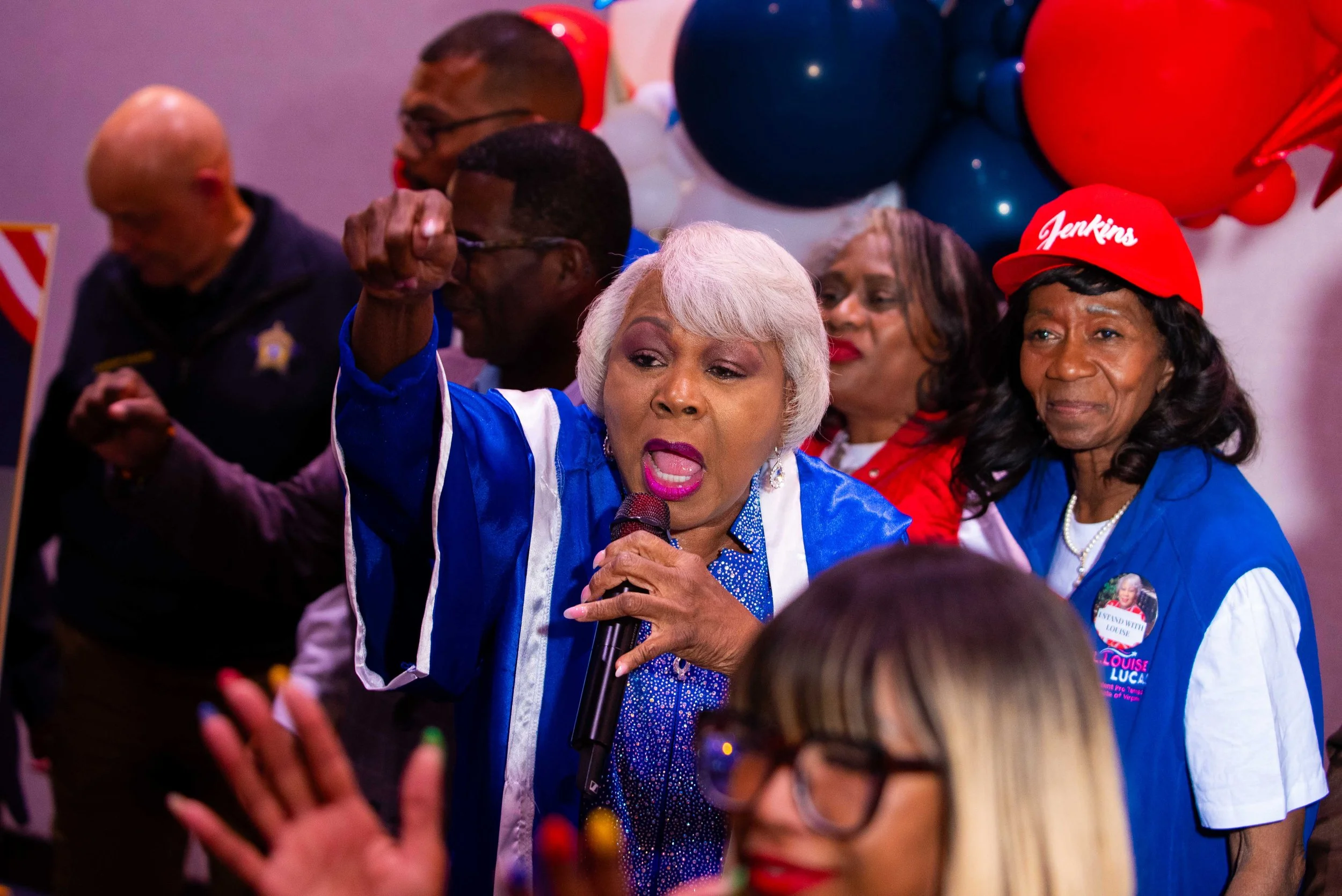 An elderly woman with blonde hair and pink lipstick, wearing a blue and white striped shirt and a blue scarf, speaking passionately into a microphone at a political rally. She is surrounded by people, some of whom are wearing red hats and shirts. Balloons and decorations are visible in the background.