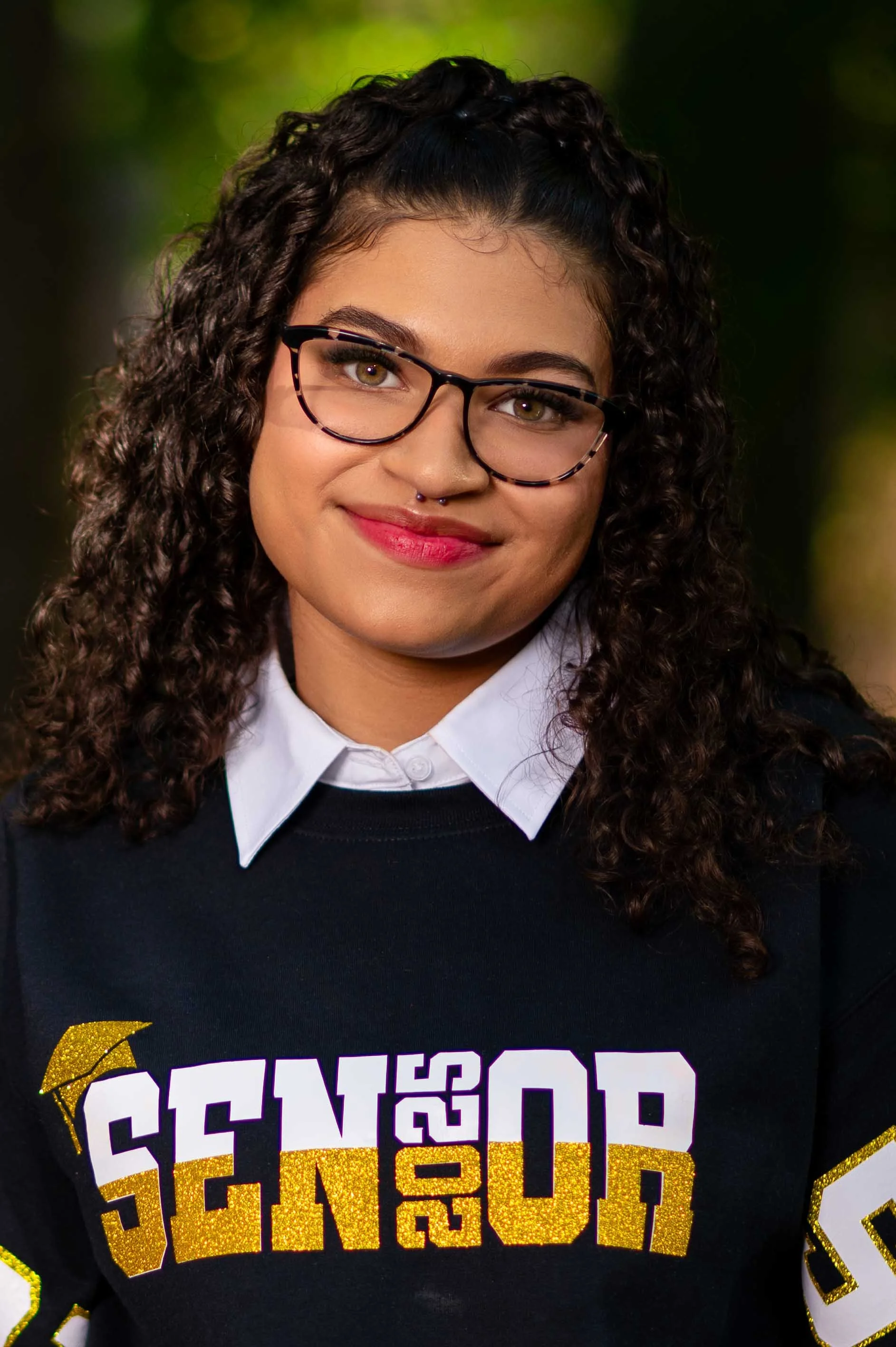 A young woman with curly dark hair, glasses, and a septum piercing, smiling outdoors, wearing a black sweatshirt with gold and white lettering that reads 'SENDER 50' and a white collared shirt underneath.