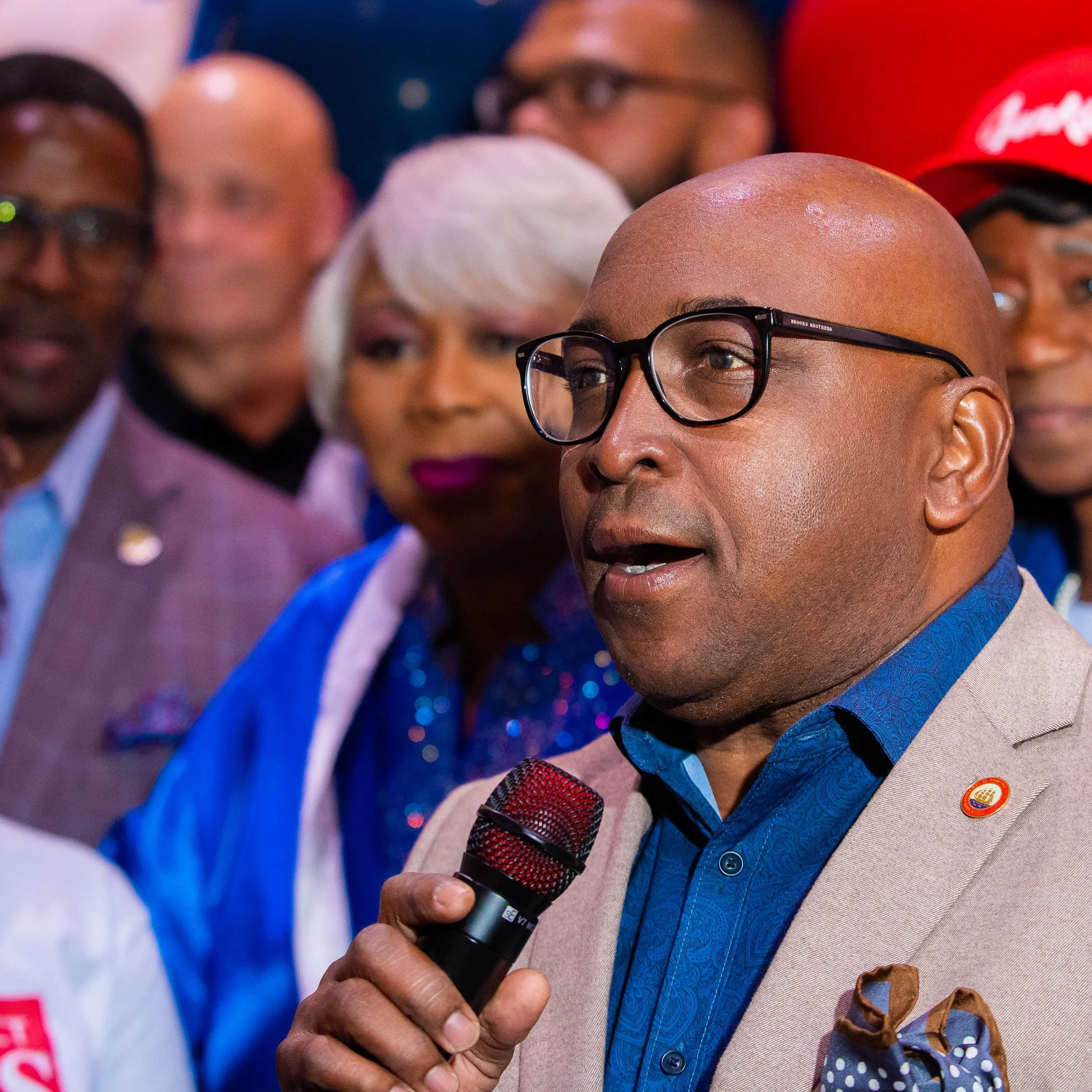Man speaking into a microphone at Rivers Casino. During an event with for Senator Lucas with a diverse group of people in the background. The