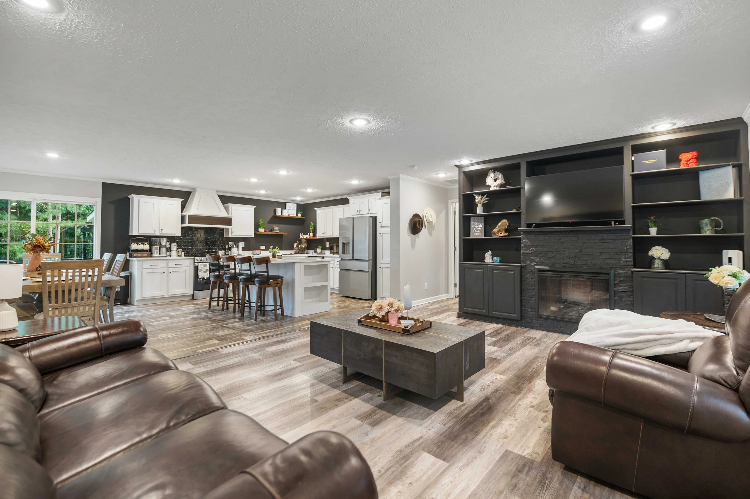 Open-concept living room and kitchen with hardwood flooring, leather sofas, a wood coffee table, a black fireplace with built-in shelving, and a kitchen with white cabinets and stainless steel appliances.