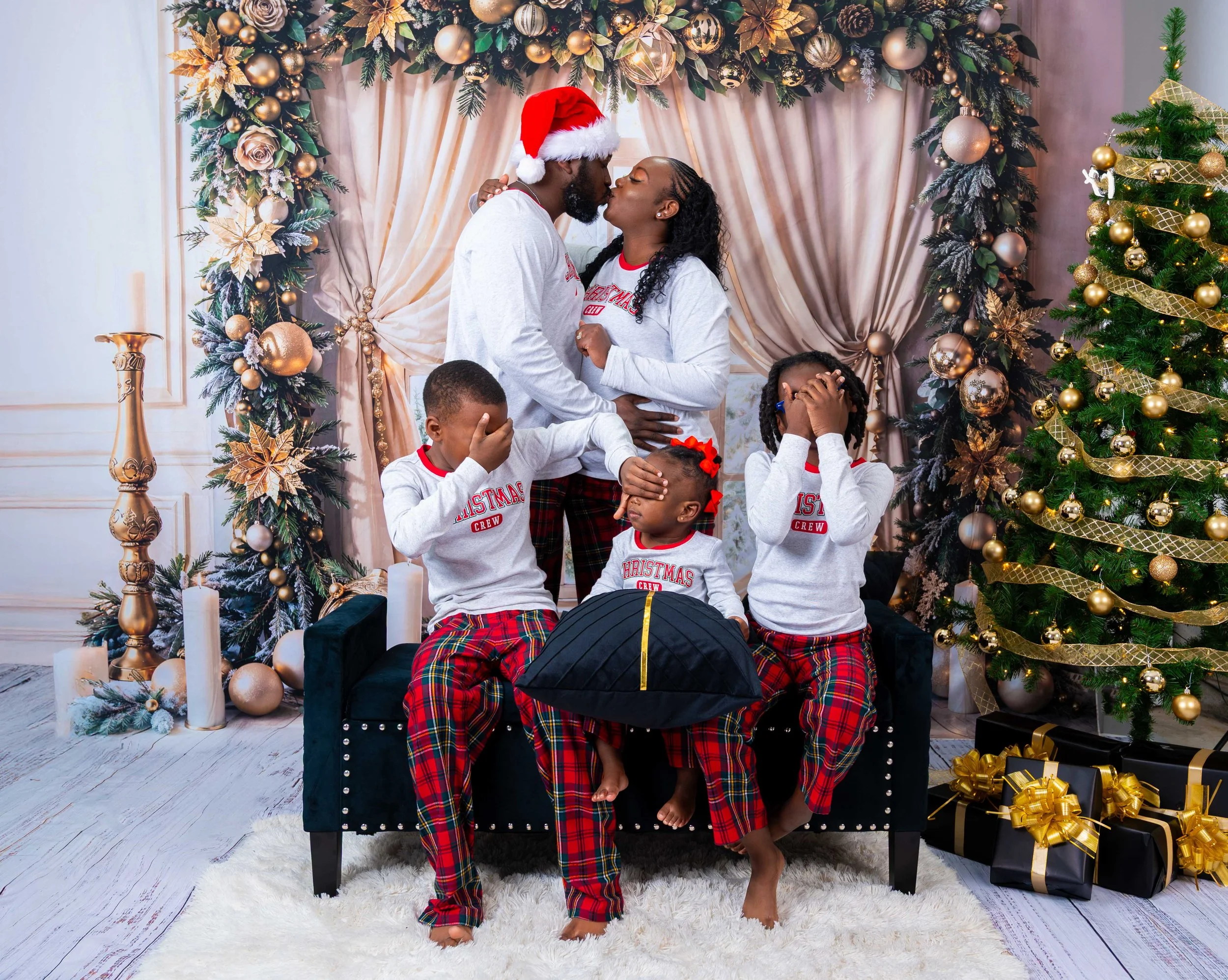 A family of six celebrating Christmas indoors. The parents are kissing, dressed in matching pajamas, with the mother wearing a Minnie Mouse shirt and father in a Santa hat. The three children, wearing matching Christmas pajamas, appear surprised or playful, covering their faces. The background features decorated Christmas trees, ornaments, and presents.