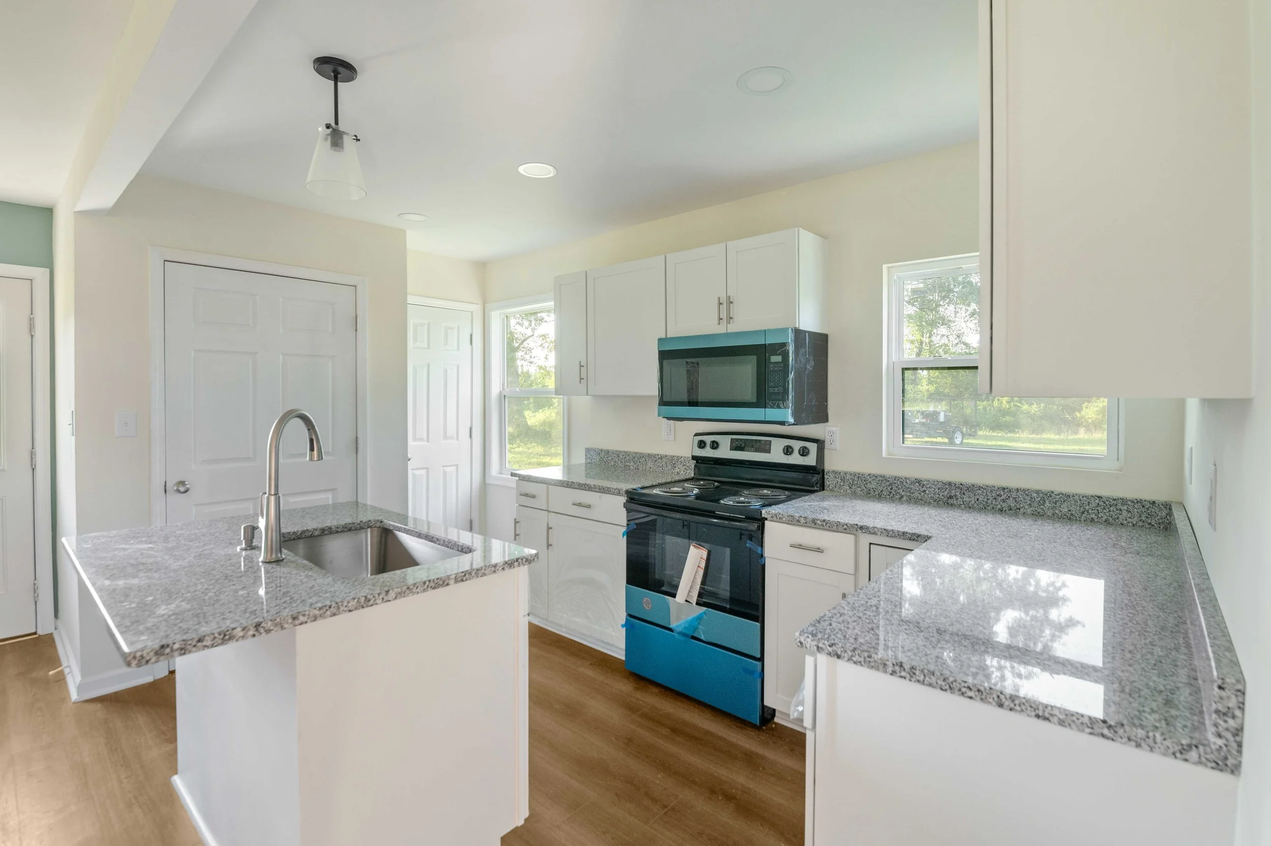 Modern kitchen with white cabinets, gray granite countertops, a black stove, microwave, and windows letting in natural light. There is an island with a sink and a silver faucet, wood flooring, and ceiling lights.