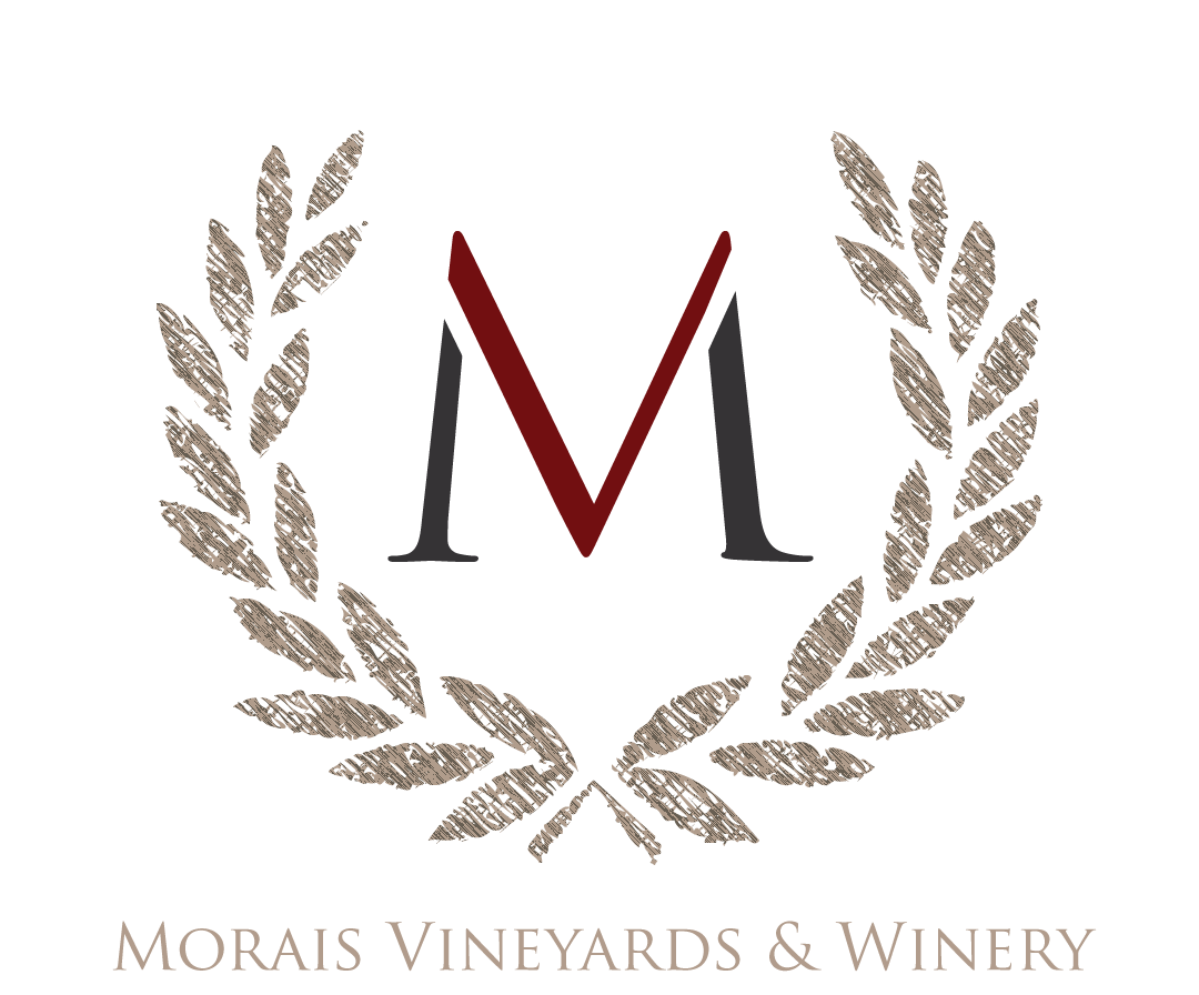 Logo of Morais Vineyards & Winery with a large black letter M, a red V in the center, surrounded by a brown laurel wreath, and the text underneath reading 'Morais Vineyards & Winery'.