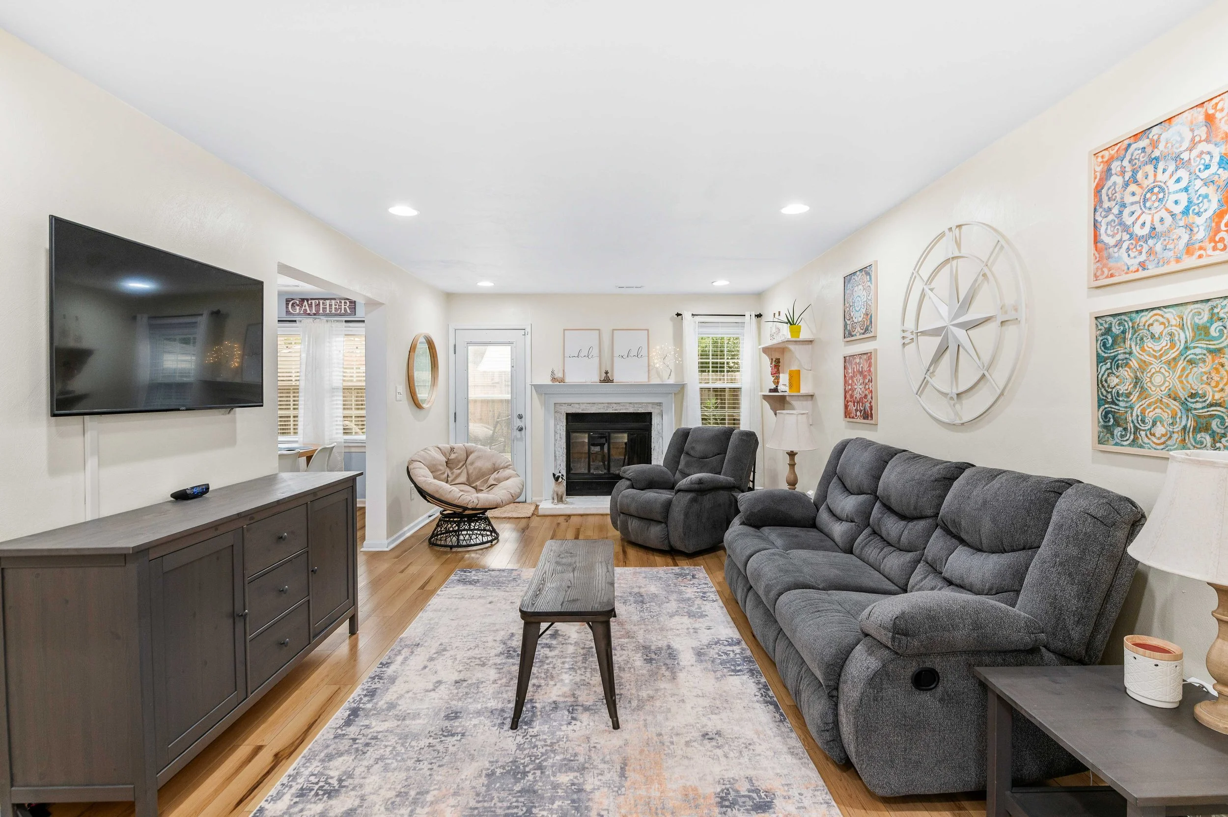 Living room with a wall-mounted flat-screen TV, a gray couch, matching recliner, and a beige papasan chair. Features artwork on the walls, a fireplace, and wooden flooring.