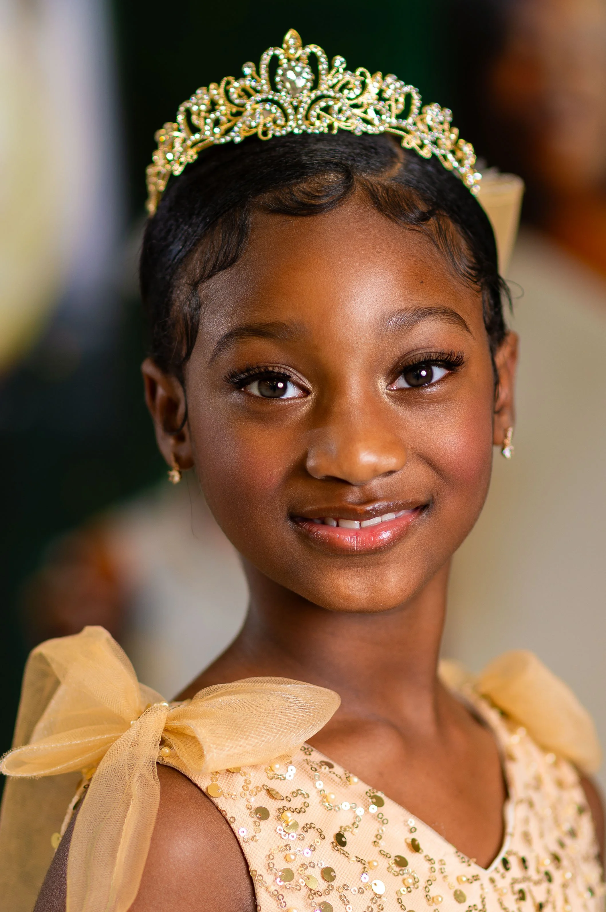 A young girl wearing a tiara and a gold sequin dress, smiling at a celebration or event.