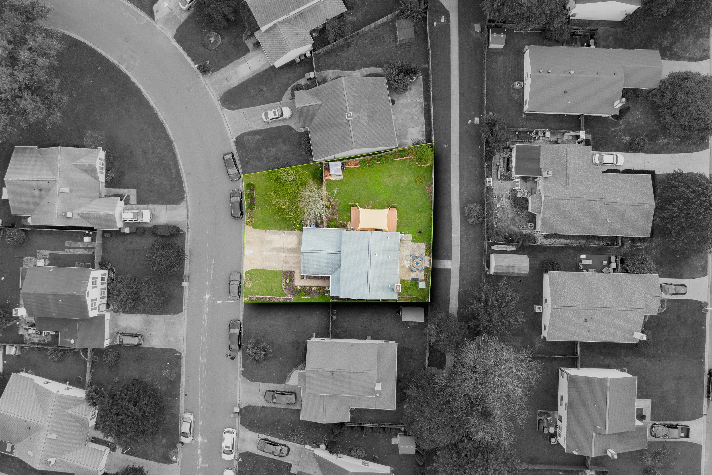 Aerial view of a house with a backyard, highlighted in color with green grass, trees, patio furniture, and a small structure, contrasting with neighboring black-and-white houses and streets.