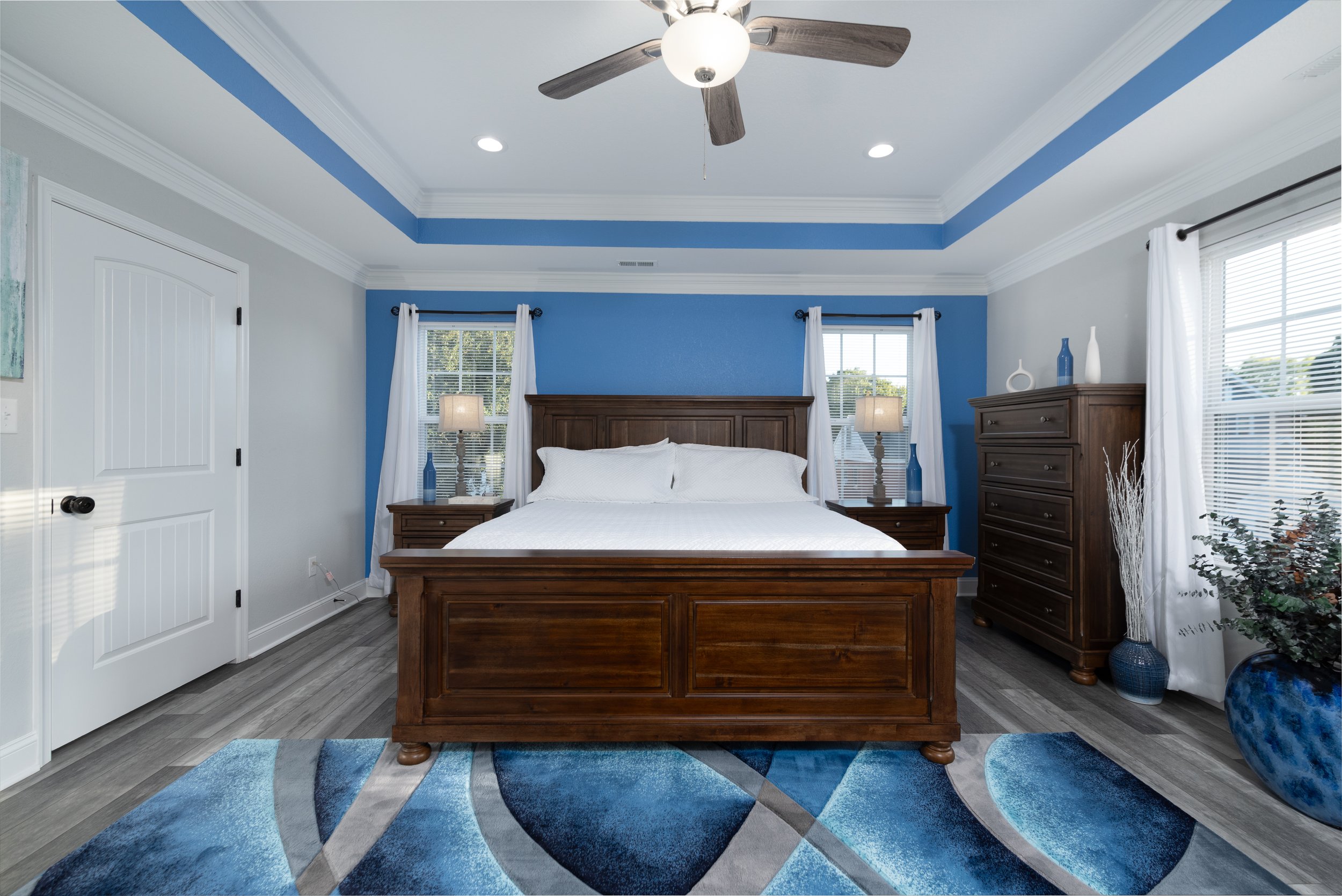 Bedroom with a wooden bed frame, white bedding, and two nightstands with lamps. Blue accent wall behind the bed, white curtains, a tall dresser, and decor vases. Ceiling fan with lights, large windows, and a patterned blue, gray, and white rug.