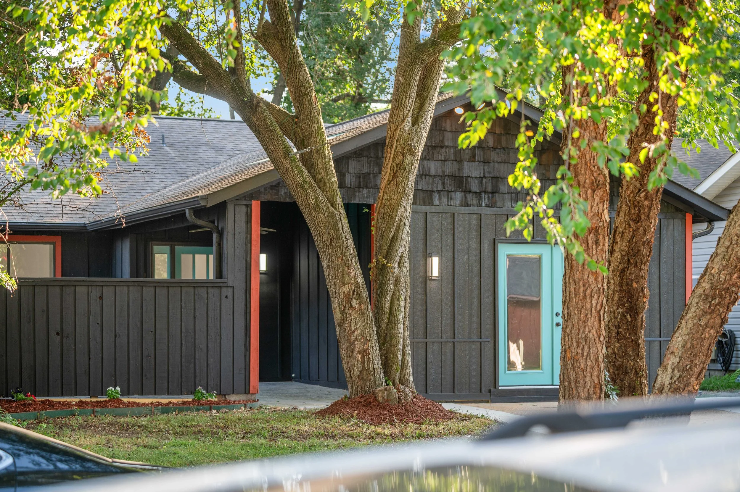 A house painted black with wooden siding, surrounded by green trees, with a teal door and a concrete pathway in front.