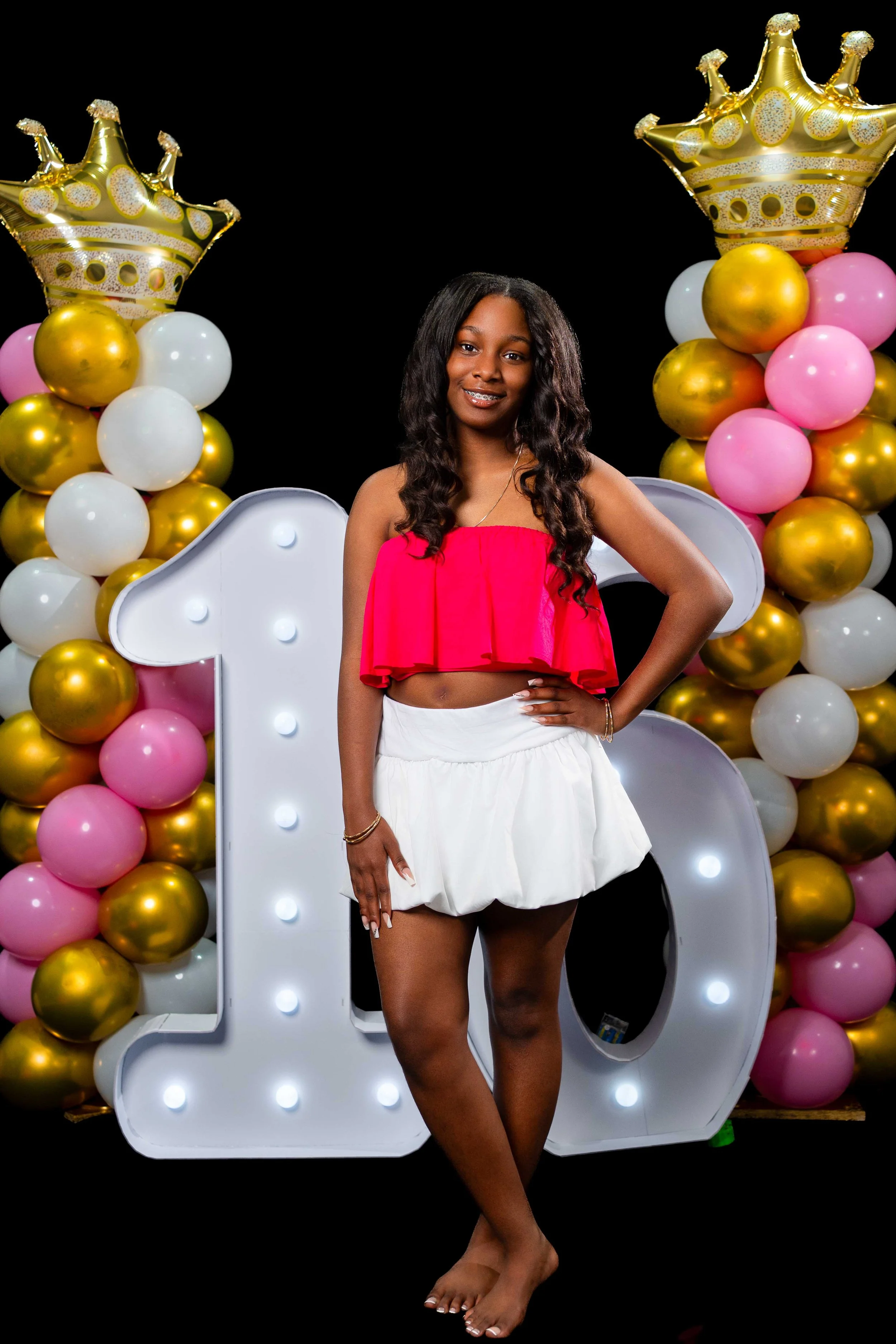 Young woman celebrating her first birthday, standing barefoot in front of large illuminated number one and balloon decorations with gold, white, and pink balloons, gold crowns, and a black background.