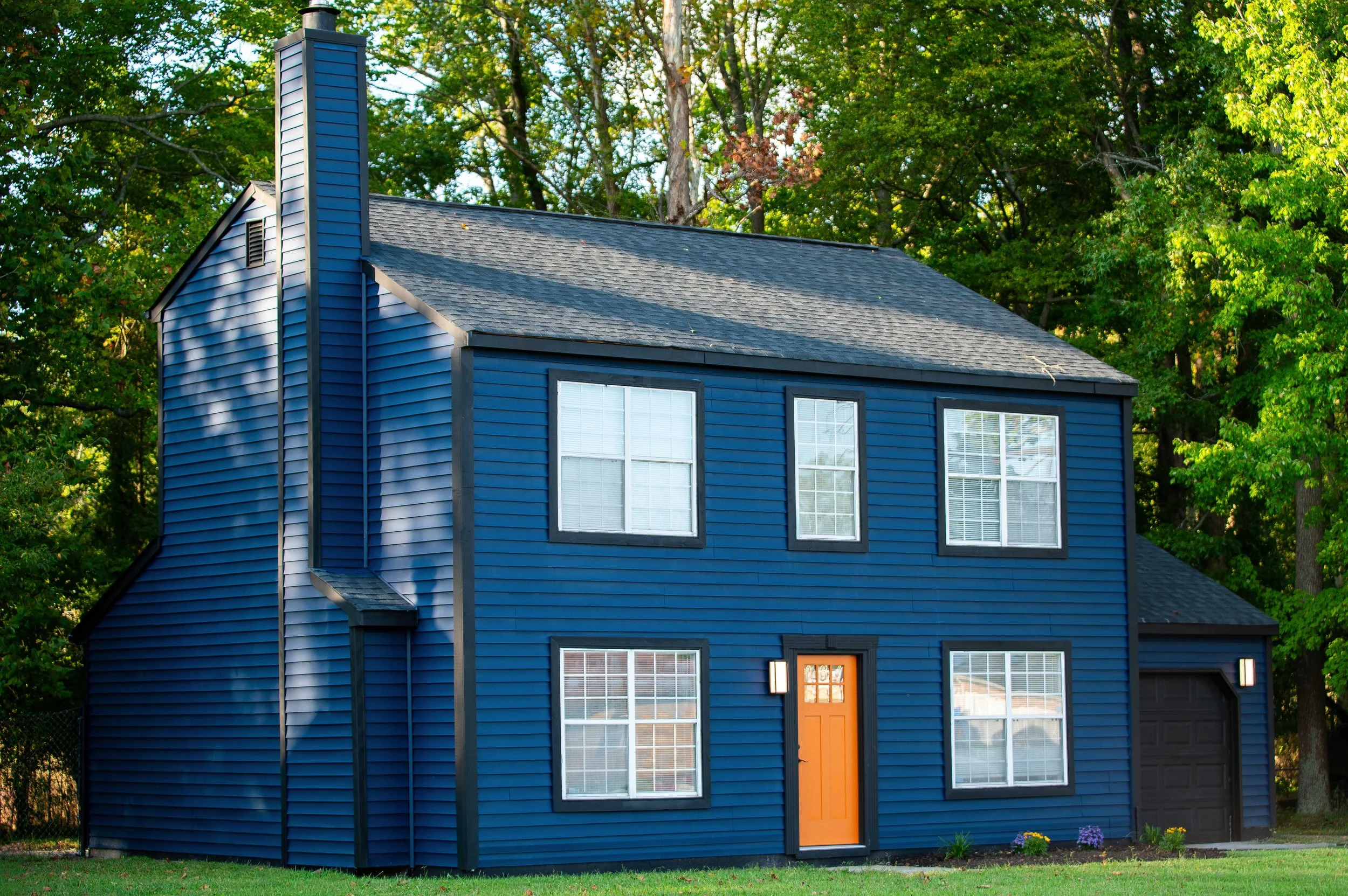 A two-story blue house with white window frames, an orange front door, and a black garage door, surrounded by green trees and grass.