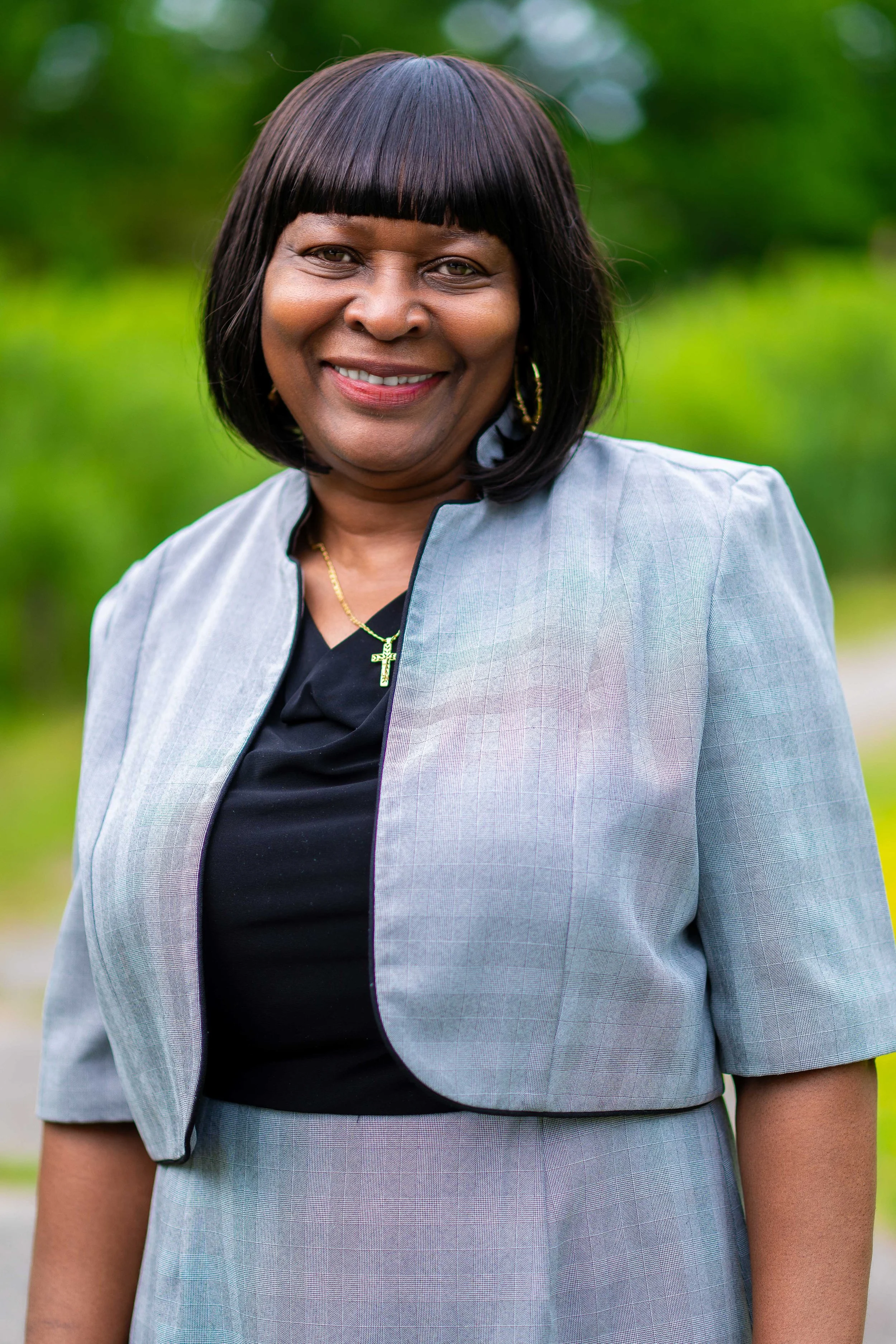 A smiling African American woman with shoulder-length black hair and bangs, wearing a light gray blazer over a black top, a gold cross necklace, and gold hoop earrings, standing outdoors with greenery in the background.