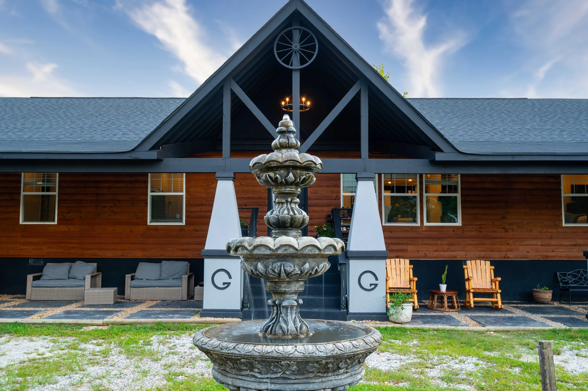 An outdoor scene with a stone fountain in front of a wooden house with a black roof and multiple windows, surrounded by outdoor seating and potted plants.