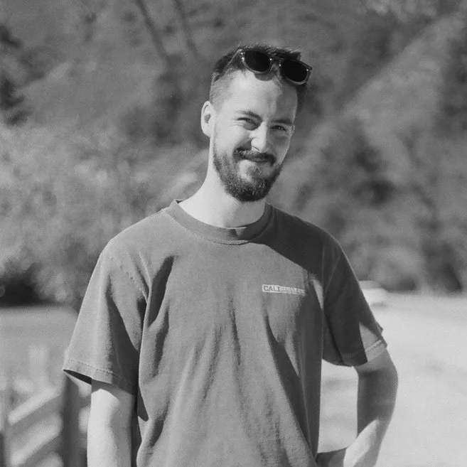 A smiling man with a beard wearing a T-shirt and sunglasses on his head outdoors.