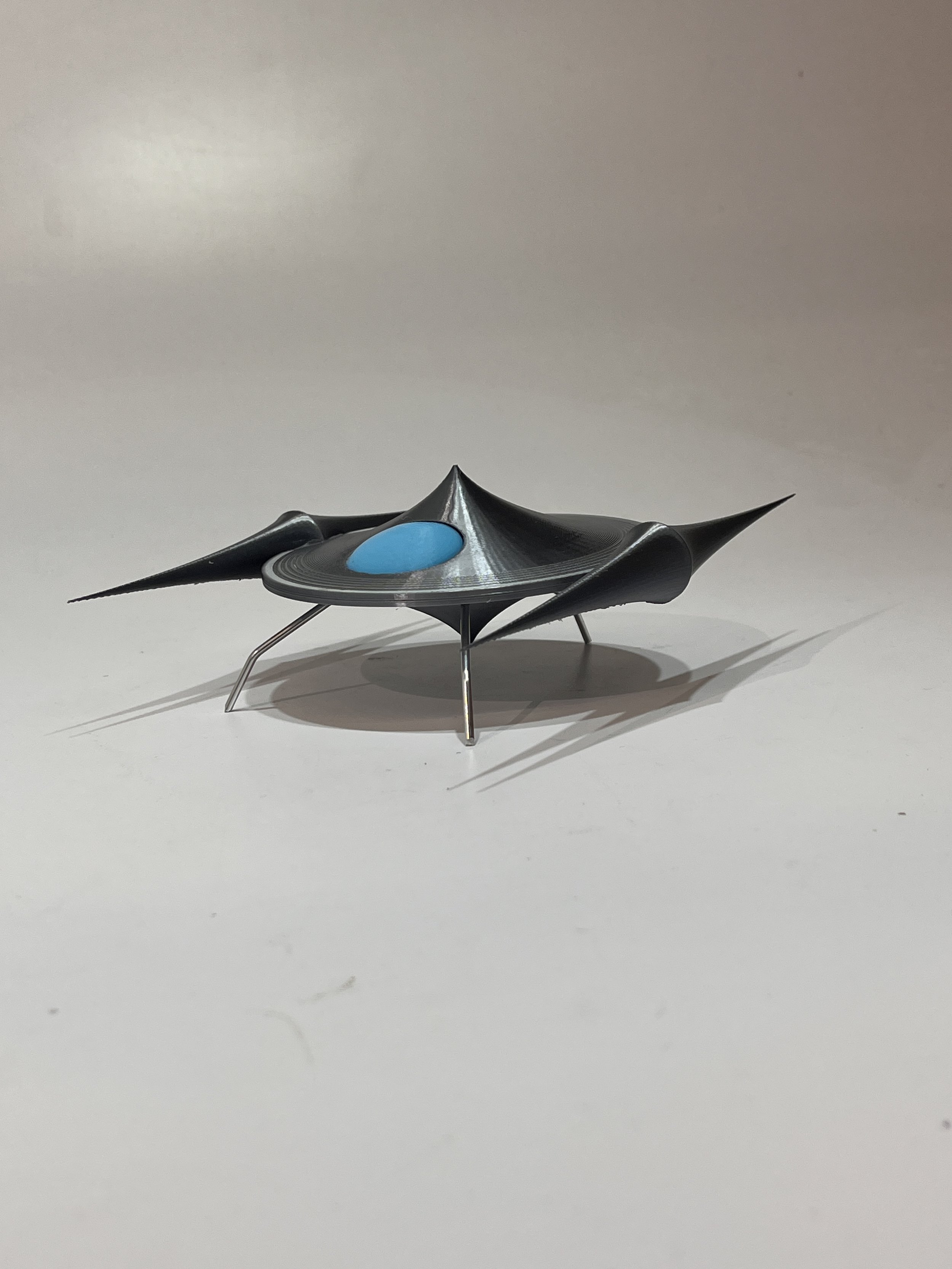 A futuristic small spacecraft with a black, sleek, aerodynamic design, three silver legs, and a glowing blue element on top, set on a white surface.