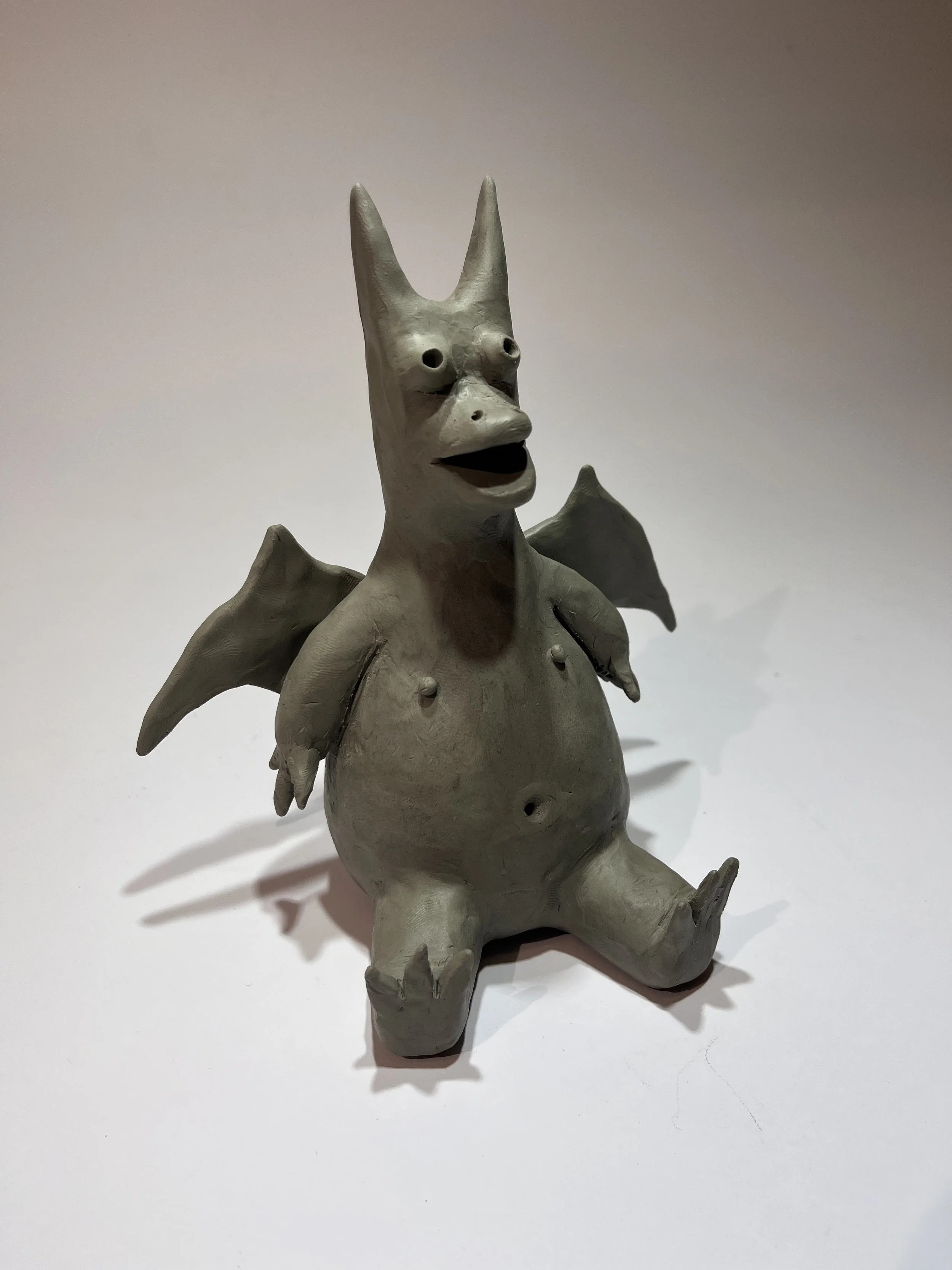 A gray, cartoonish figure with a humanoid body, bat-like wings, and an animal-like face with pointed ears and a large mouth, made from a smooth material, sitting on a plain surface.