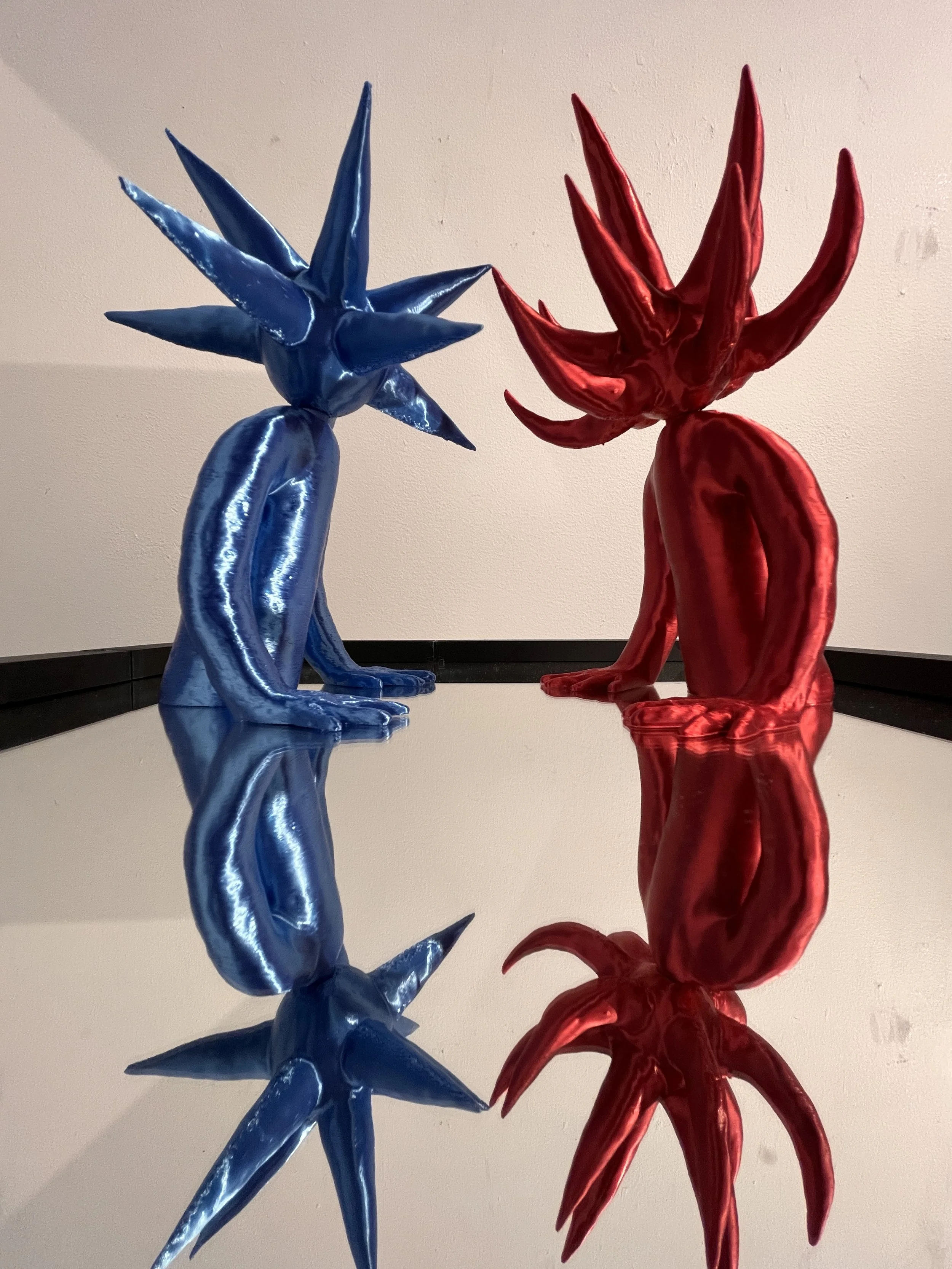 Artistic sculptures of a blue spiky figure and a red spiky figure, with both figures reflecting on a mirrored surface.