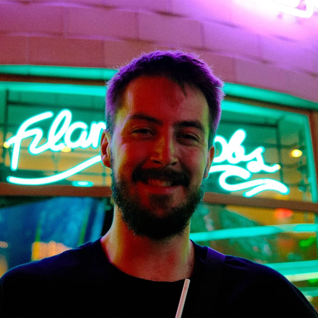 A man with a dark beard smiling in front of a neon sign that says "flamingos" at night.