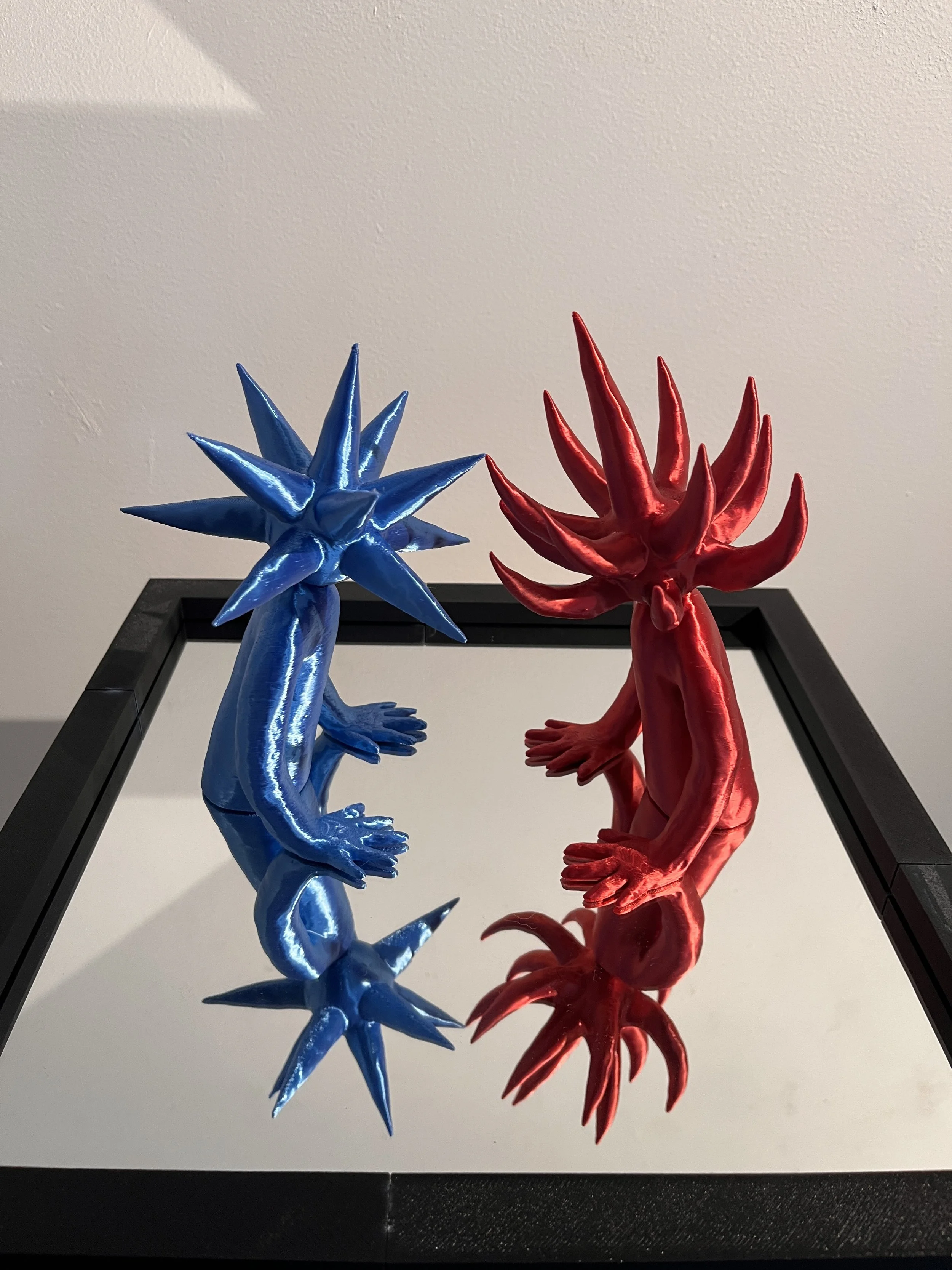 Two sculptures resembling alien or fantasy creatures, one blue and one red, with spiky hair and elongated limbs, are positioned to face each other on a reflective surface.